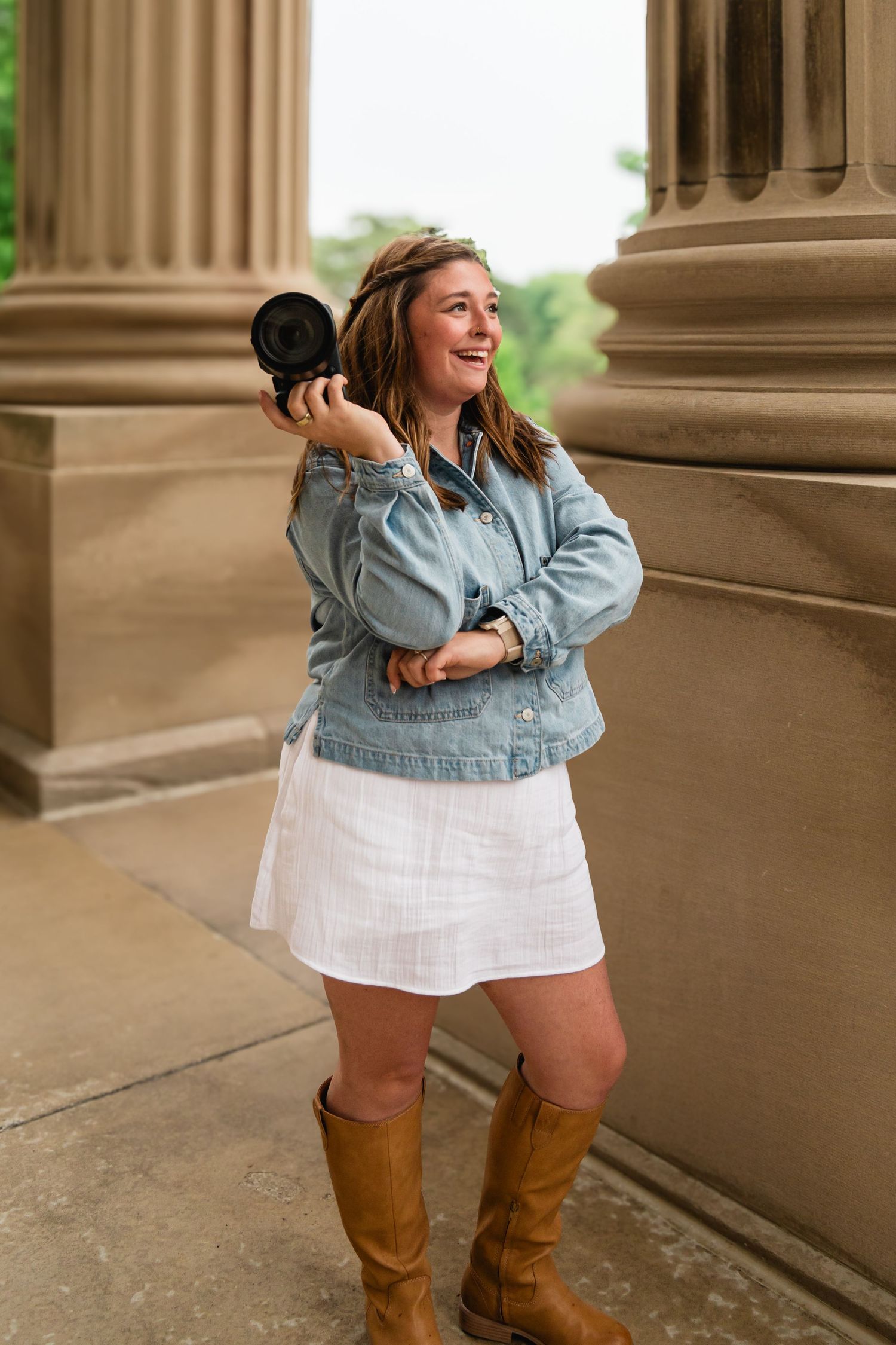 Smiling female photographer holding a camera beside stone columns in a denim jacket and white skirt.