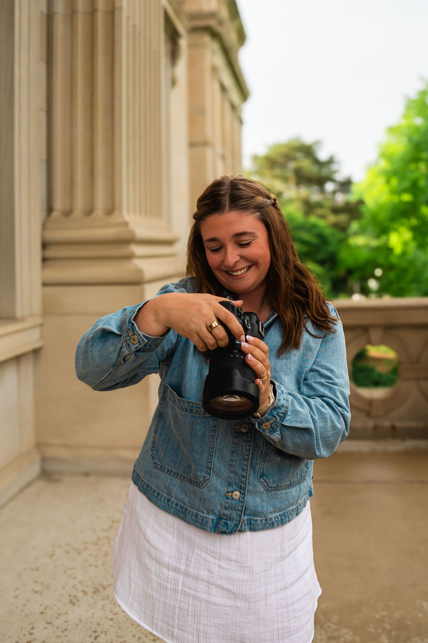A smiling young woman in a denim jacket and white skirt reviews photos on her camera outdoors near stone columns.