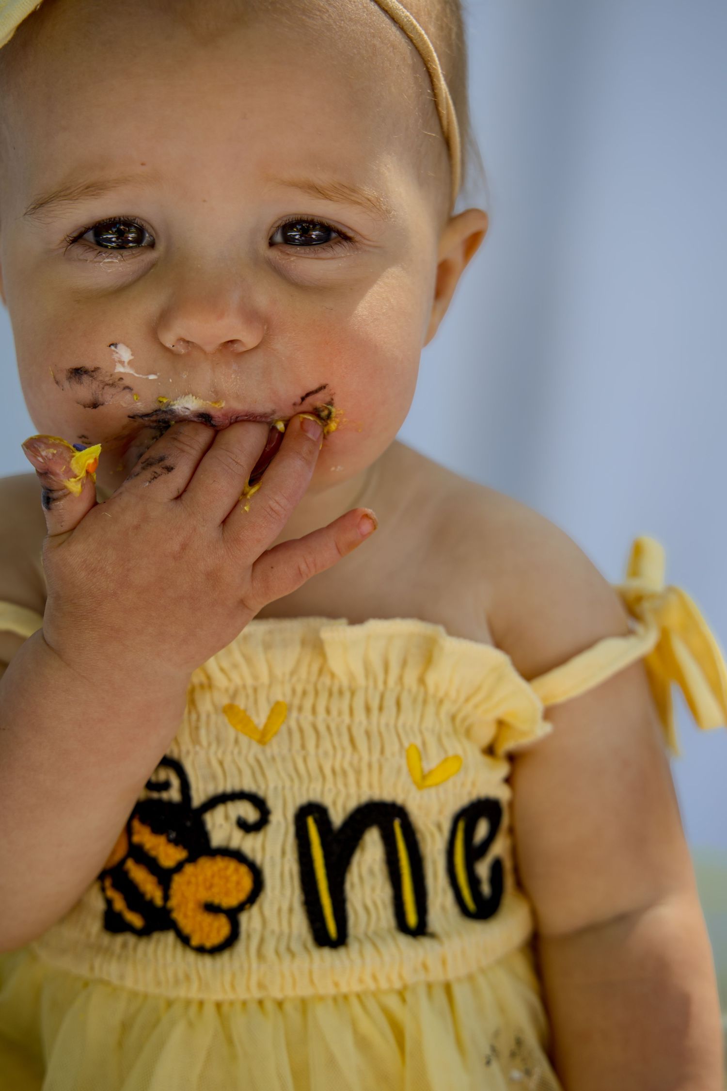 Baby girl in yellow bee dress eating birthday cake with messy hands and face during first birthday smash cake session.