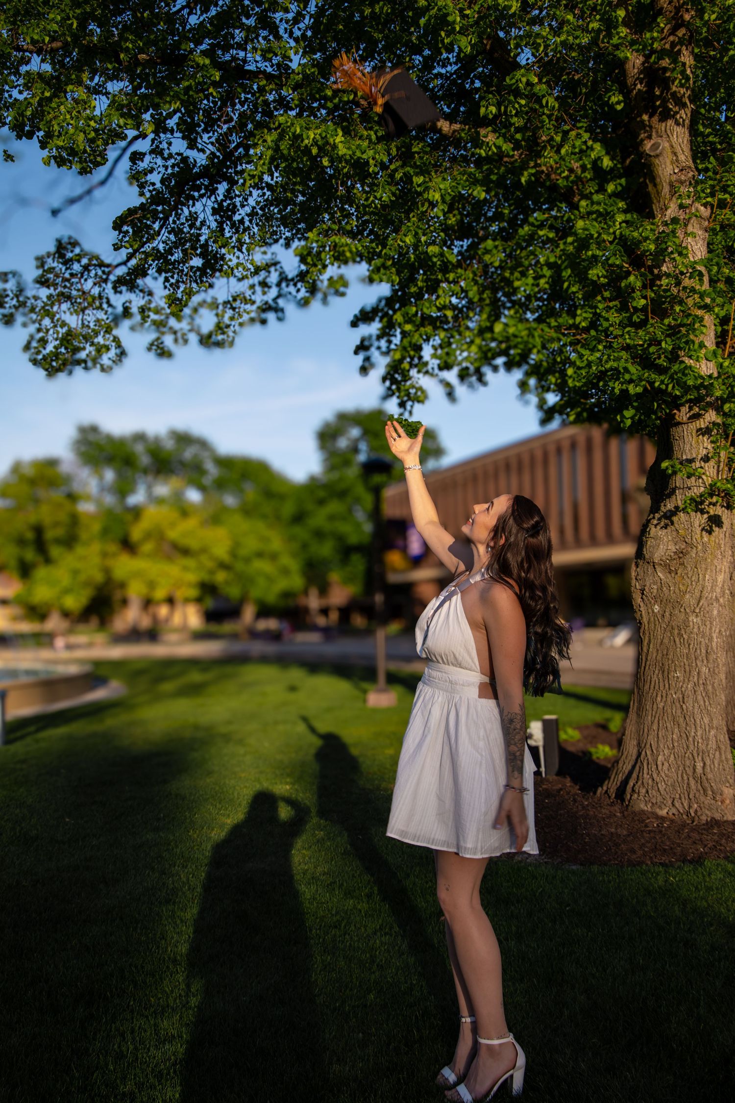 Young woman in white dress tossing graduation cap under a tree on sunny campus.