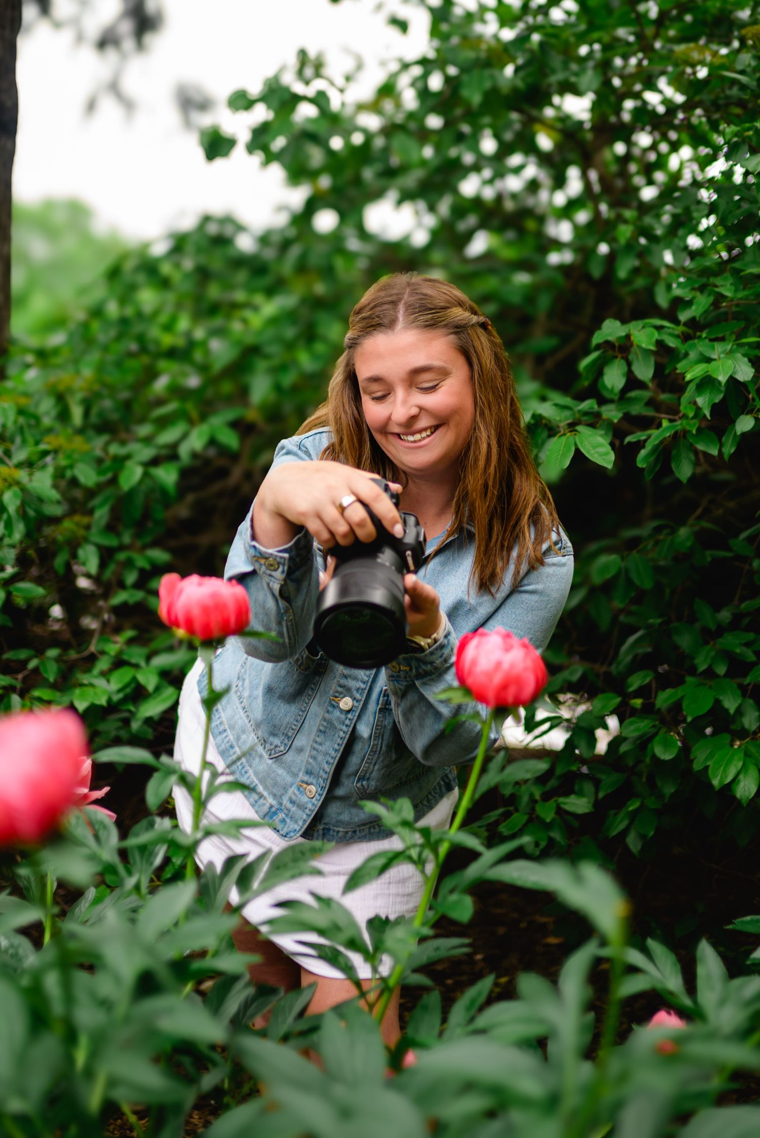 Female photographer smiling while photographing pink peony flowers in a lush green garden setting.