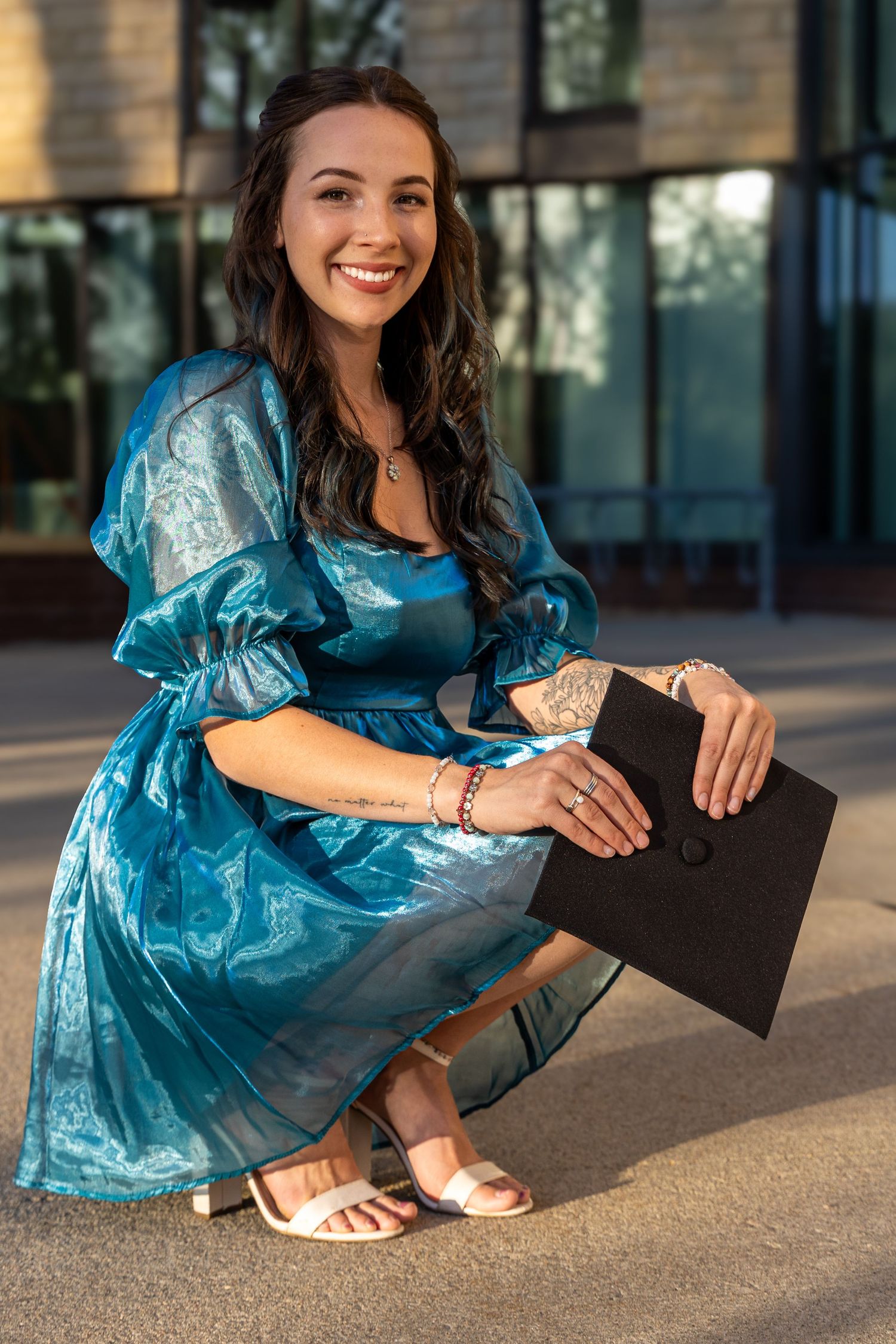 Smiling brunette woman in a teal dress crouching outdoors holding a graduation cap.