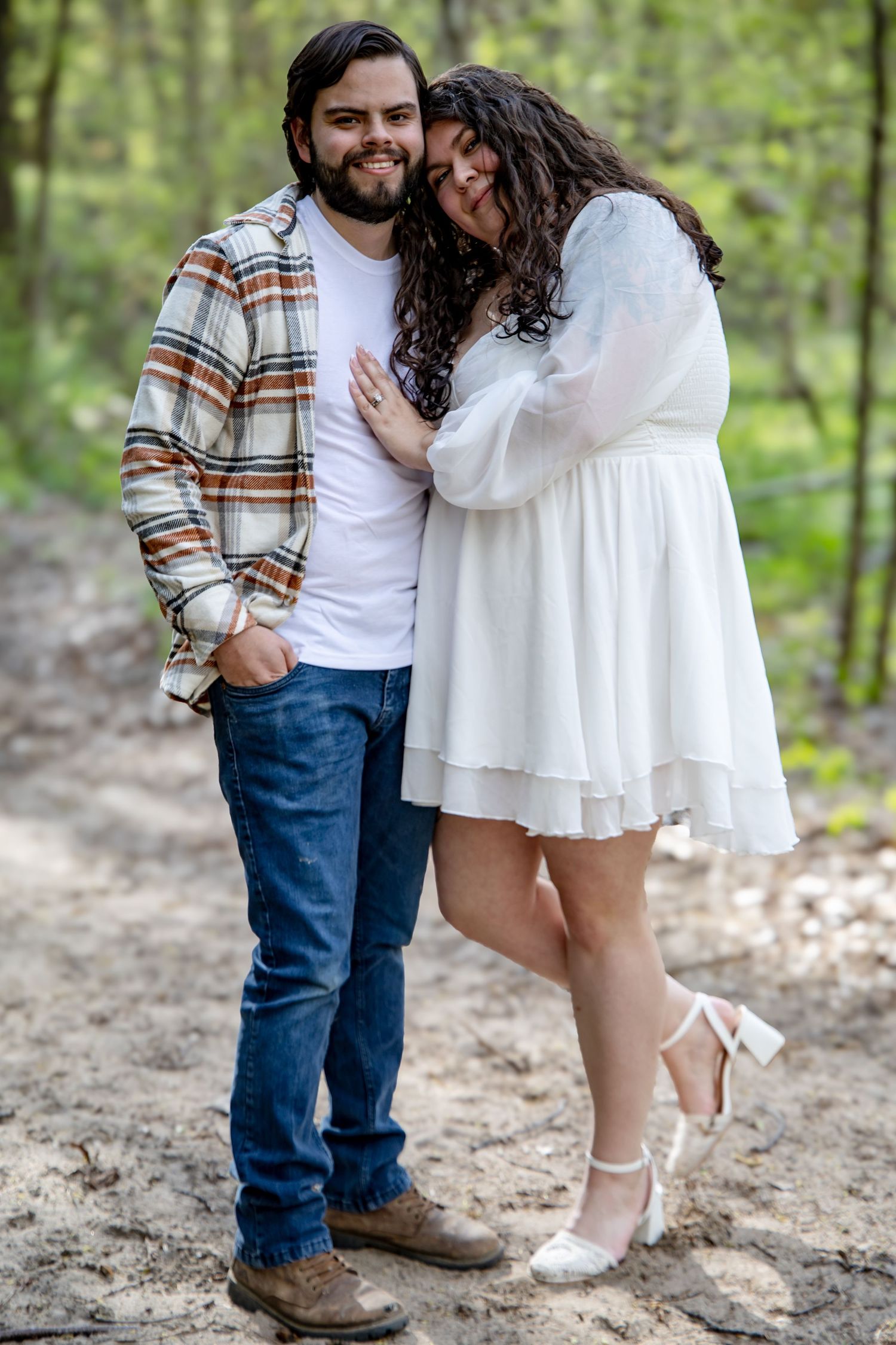 Couple embraces romantically in a forest, woman in white dress leans on smiling bearded man.