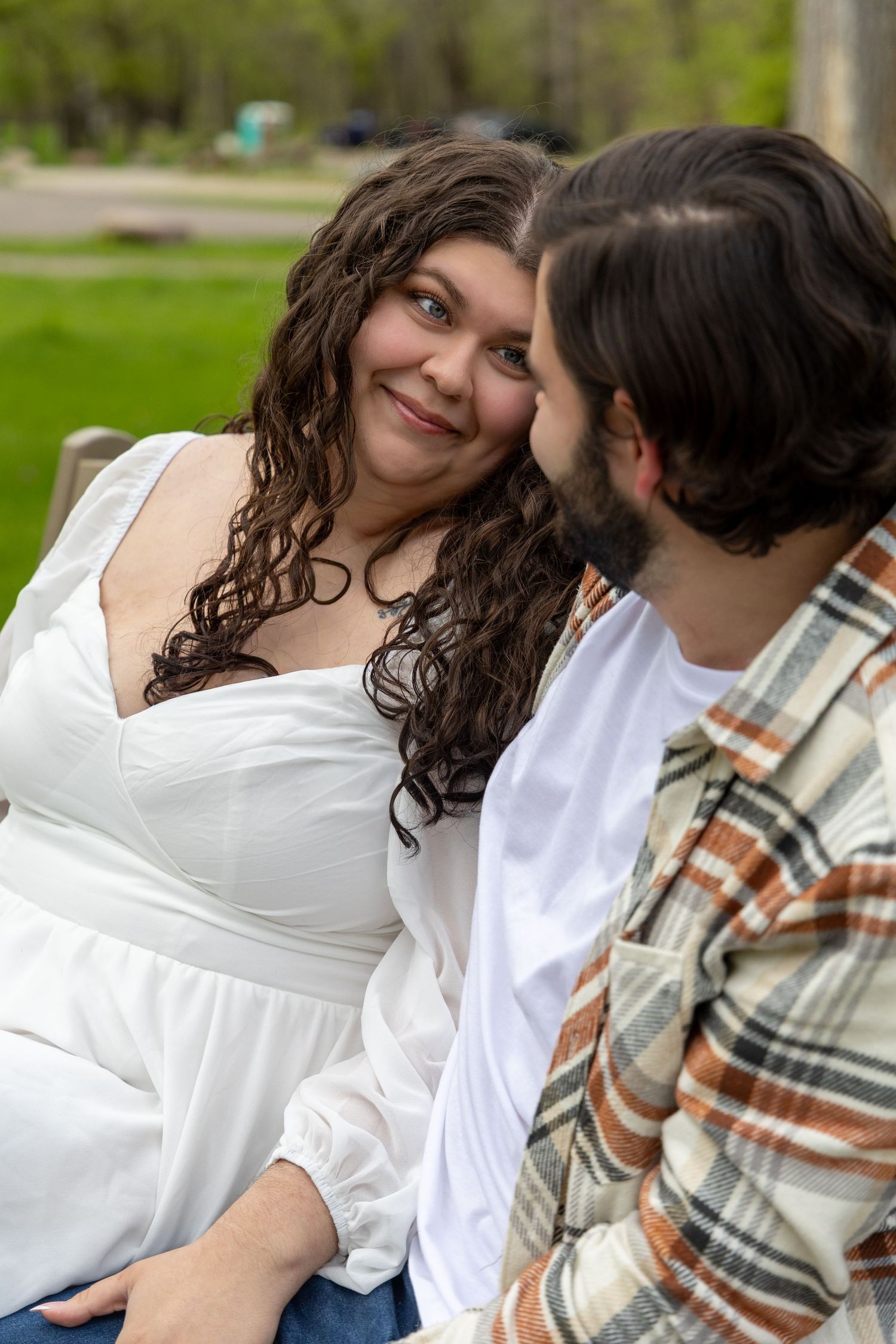 Smiling curly-haired woman in white dress being kissed on cheek by bearded man in park.