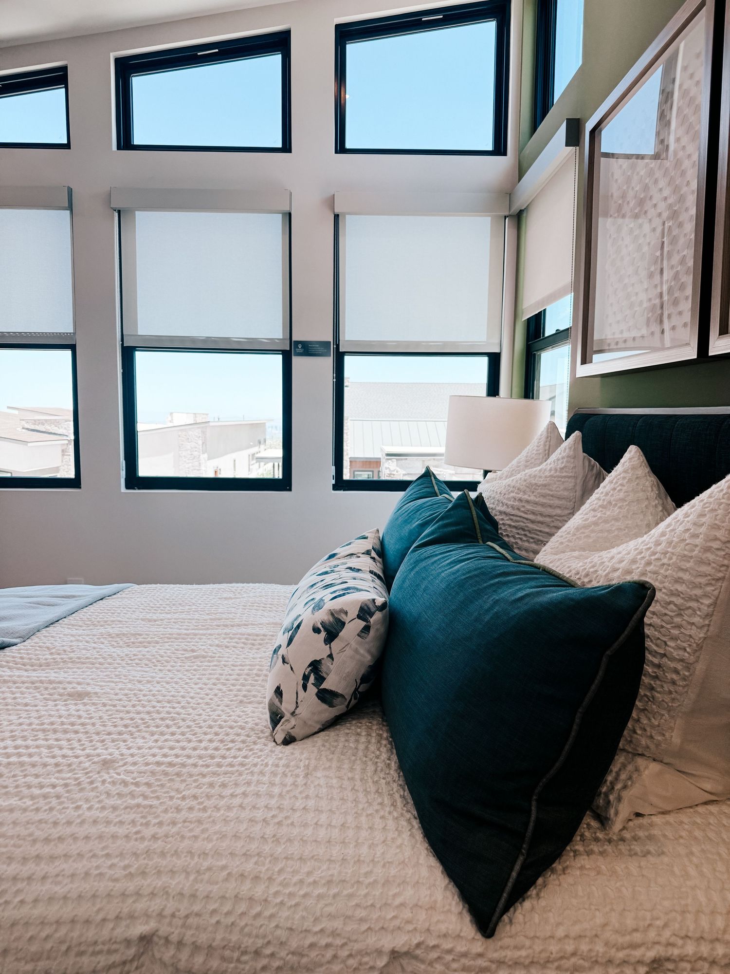 Modern bedroom with teal velvet pillows on white bedding, large windows with roller shades overlooking city rooftops.