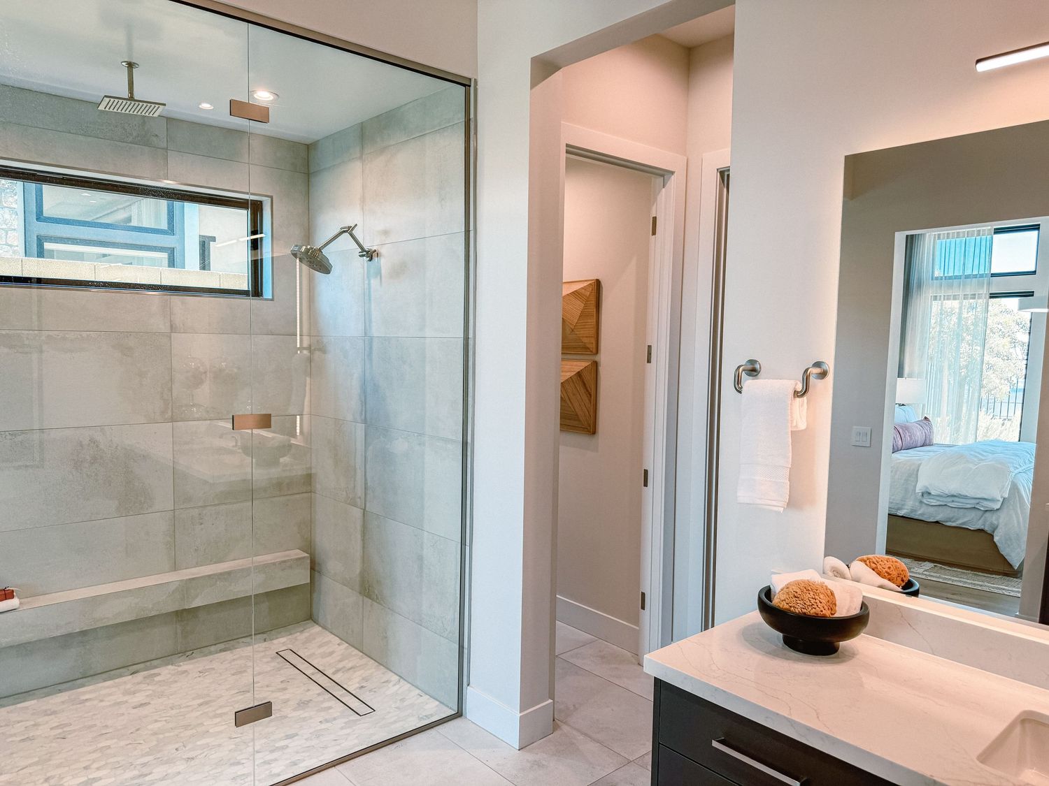 Modern bathroom with glass walk-in shower, marble tile, dark vanity, and a view into a bright bedroom.