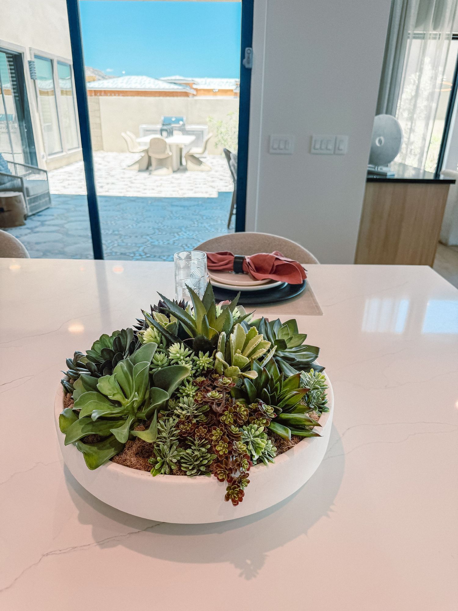 White bowl with lush succulent arrangement on marble countertop, with outdoor patio seating visible through glass doors.