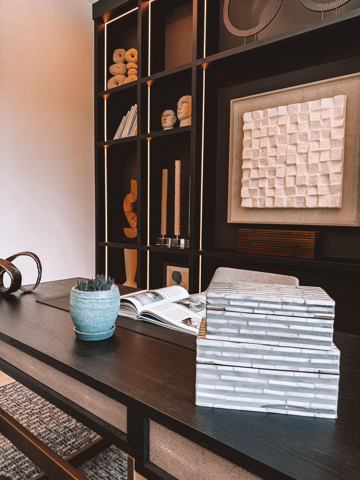 Dark wood bookshelf with decorative objects and artwork above a console table with stacked books and a blue ceramic pot.