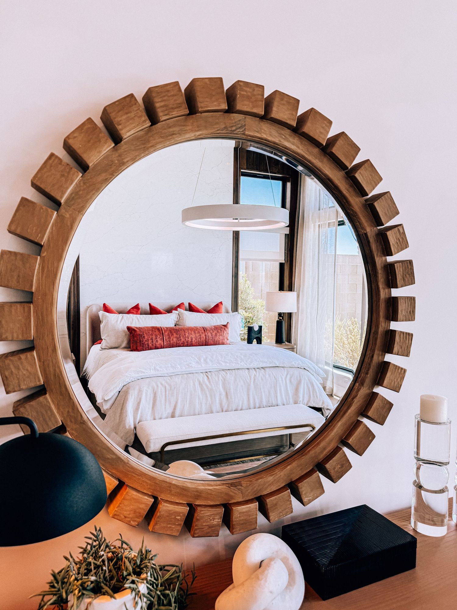 Round wooden sunburst mirror reflecting a cozy bedroom with white bedding, red pillows, and a pendant light.