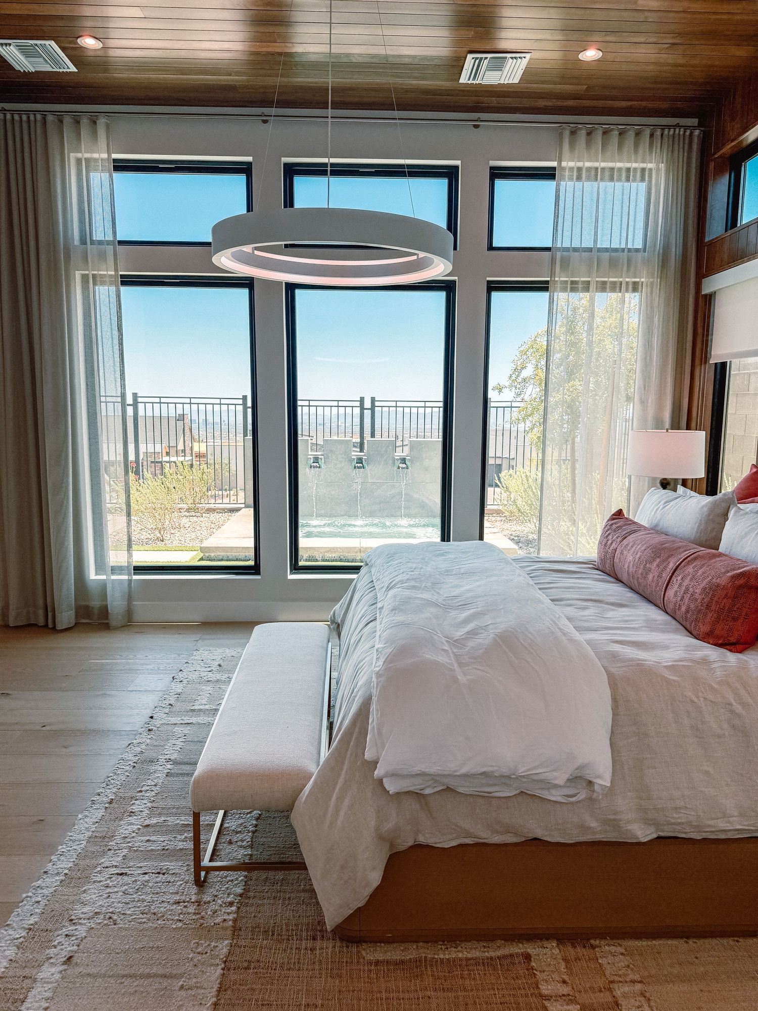Modern bedroom with circular pendant light, large windows overlooking a scenic view, white bedding, and hardwood floors.