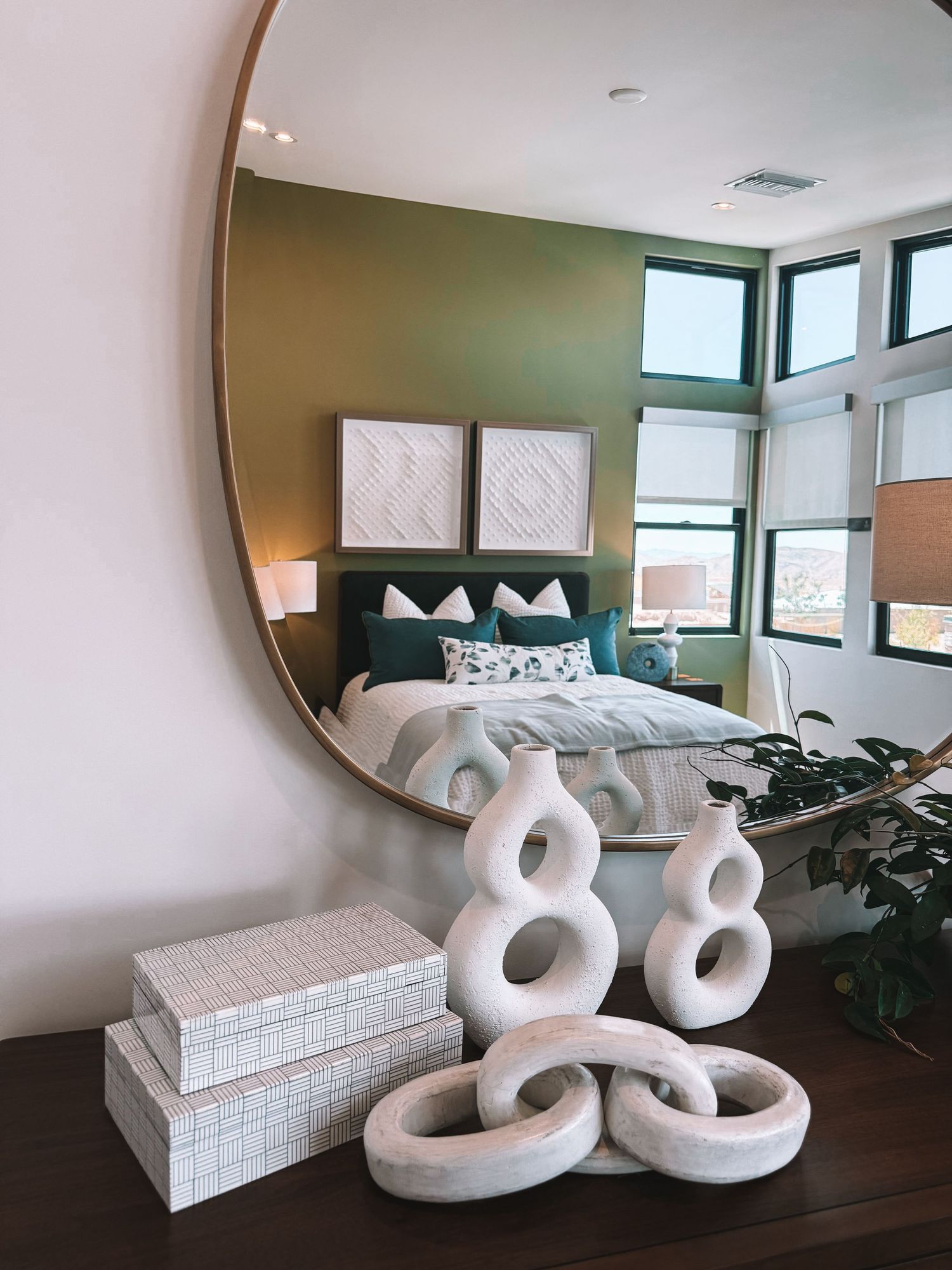 Modern bedroom with round gold mirror, white sculptural vases, decorative boxes on dresser, and green accent wall.