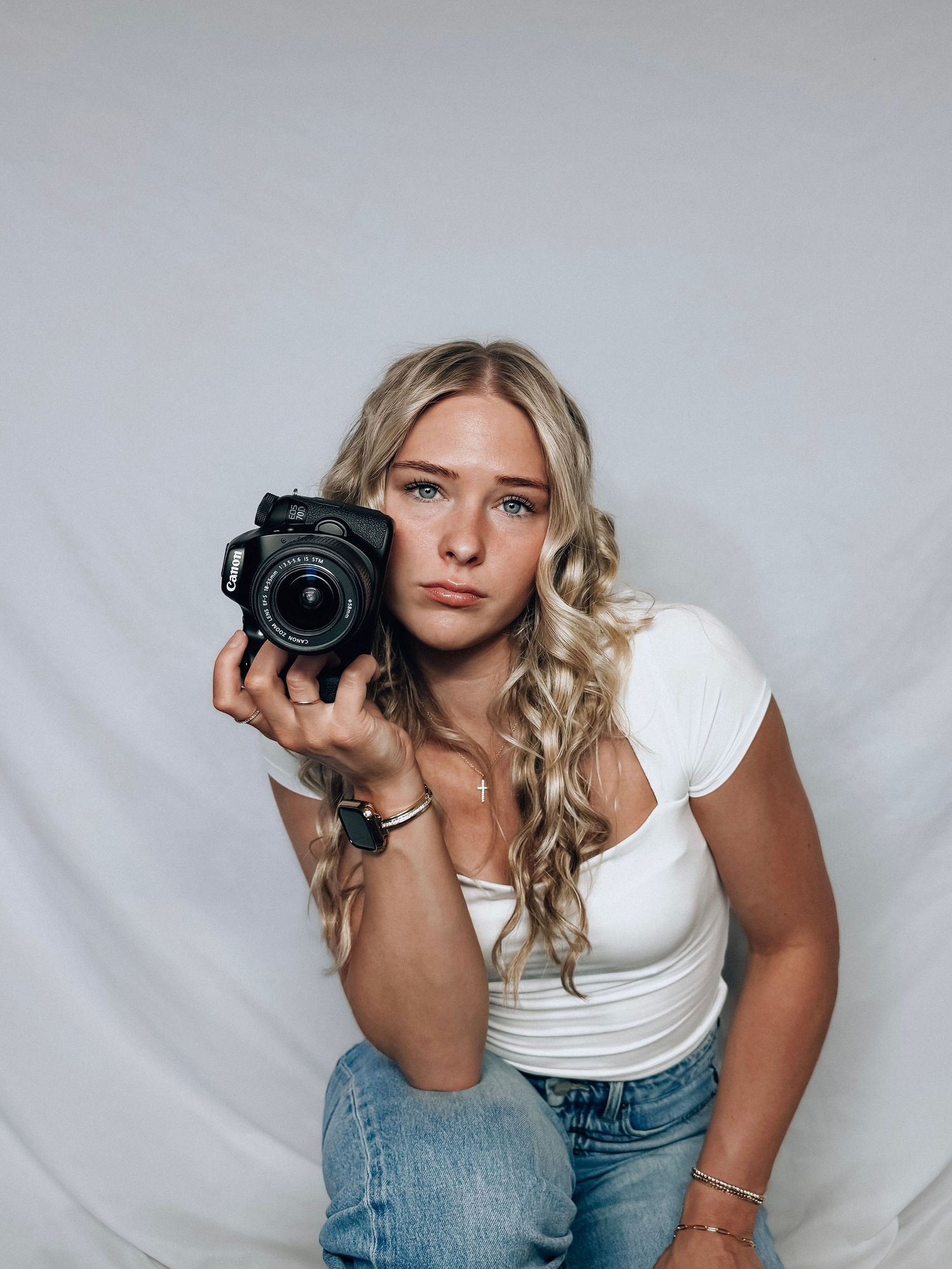 Young blonde woman holding a DSLR camera in a white studio setting, wearing a white crop top and jeans.