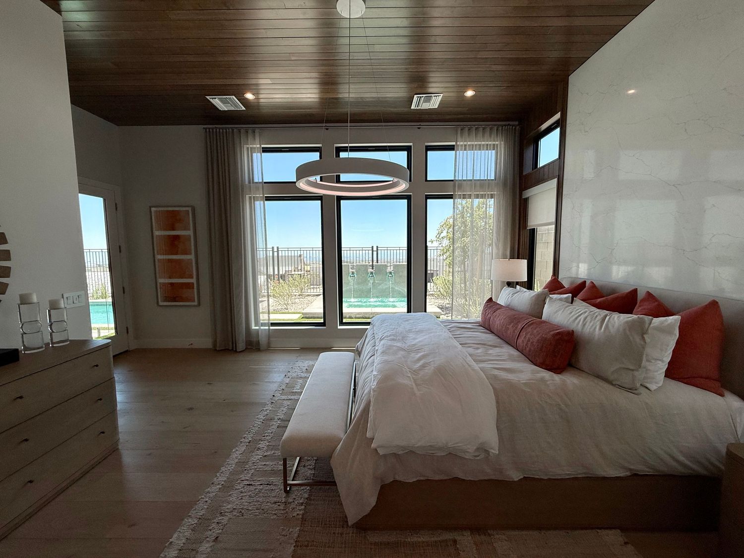 Luxury master bedroom with wood ceiling, modern chandelier, large windows, hardwood floors, and king bed with coral accent pillows.