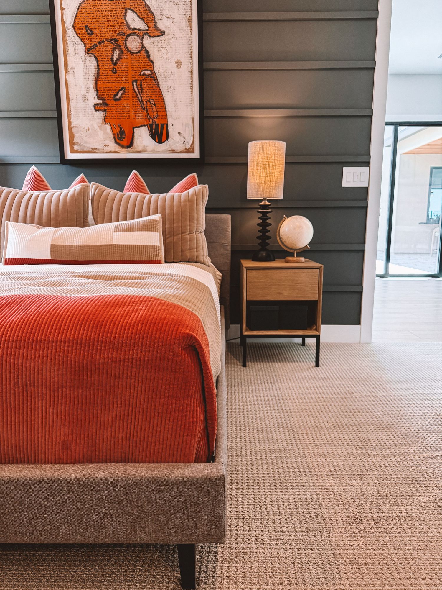 Modern bedroom with orange bedding, decorative pillows, wood nightstand, globe lamp, and abstract wall art on dark shiplap wall.
