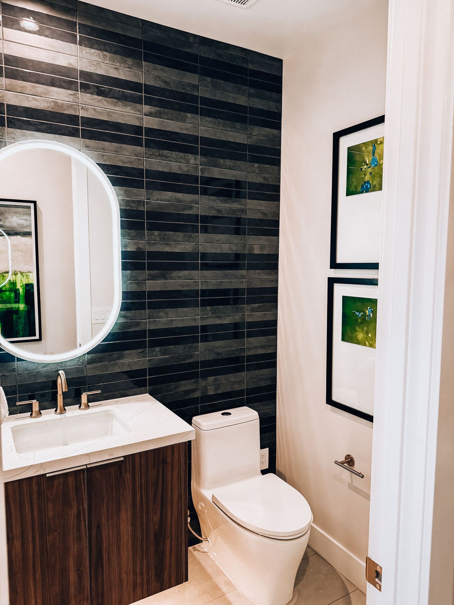 Modern bathroom with dark wood-look tile accent wall, round LED mirror, walnut vanity, white toilet, and green botanical framed art.