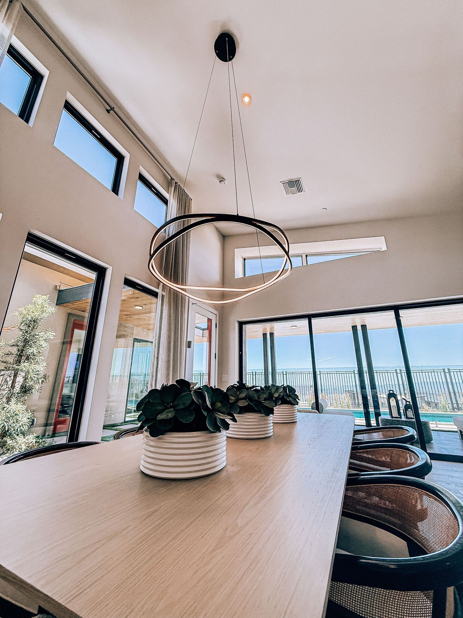 Modern dining room with circular pendant light, wooden table, white planters, and floor-to-ceiling windows overlooking a pool.