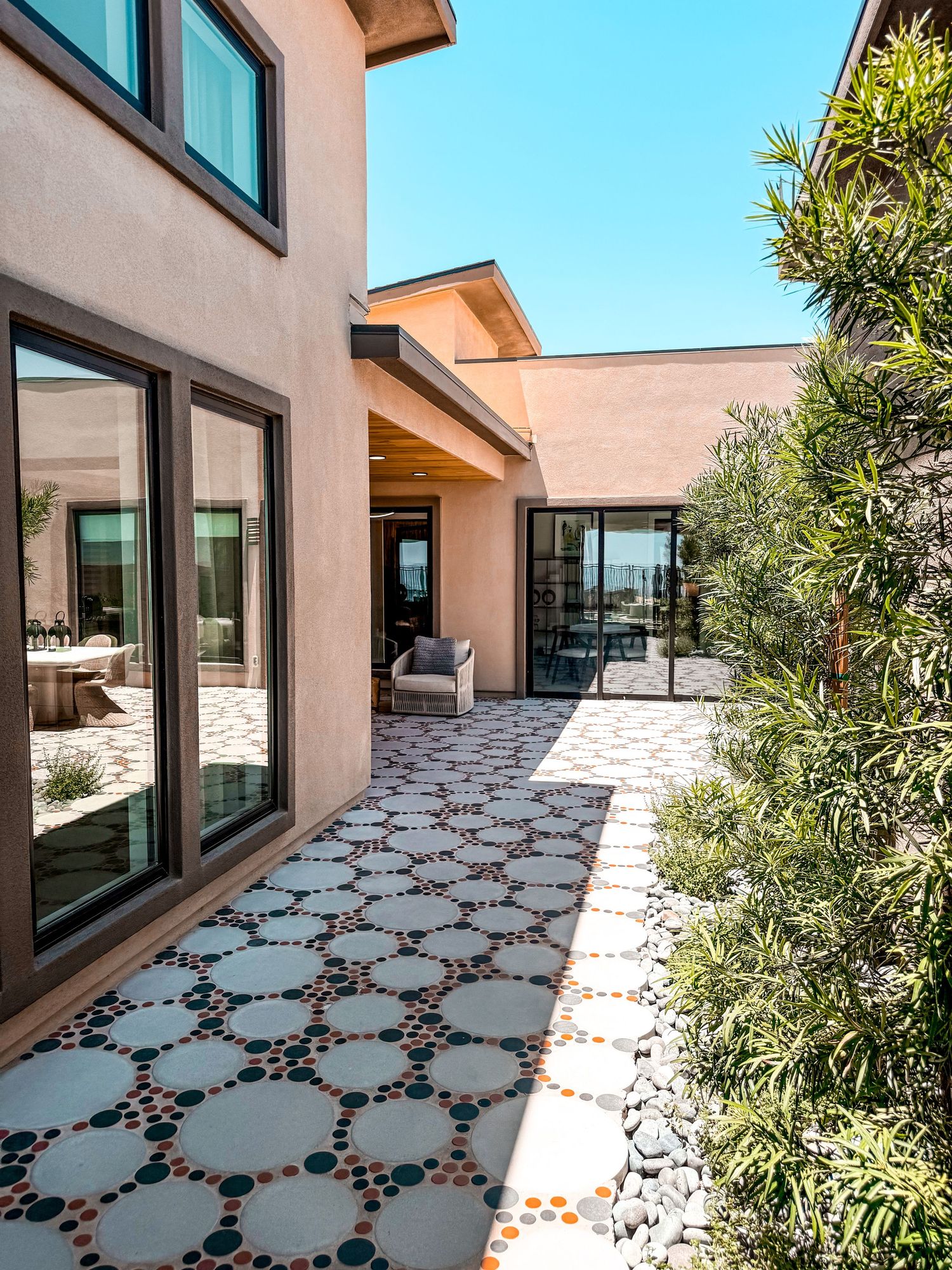 Modern home courtyard with decorative mosaic tile pathway, large glass doors, and lush greenery under a clear blue sky.
