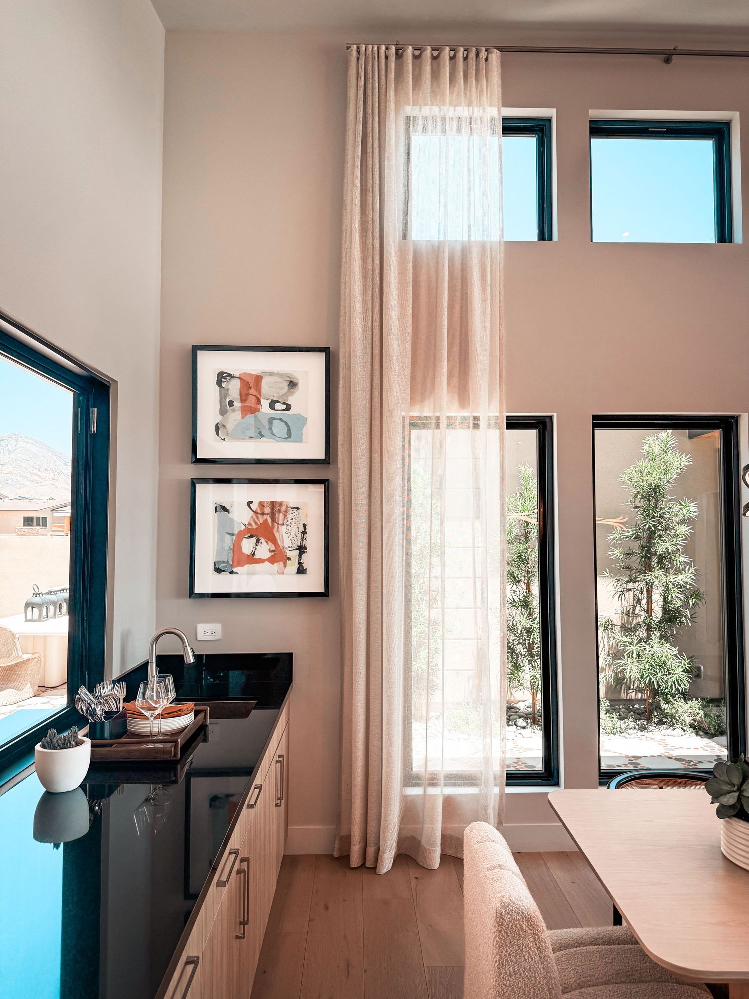 Modern kitchen nook with sheer curtains, framed wall art, dark countertop bar, and large windows overlooking a sunny patio.