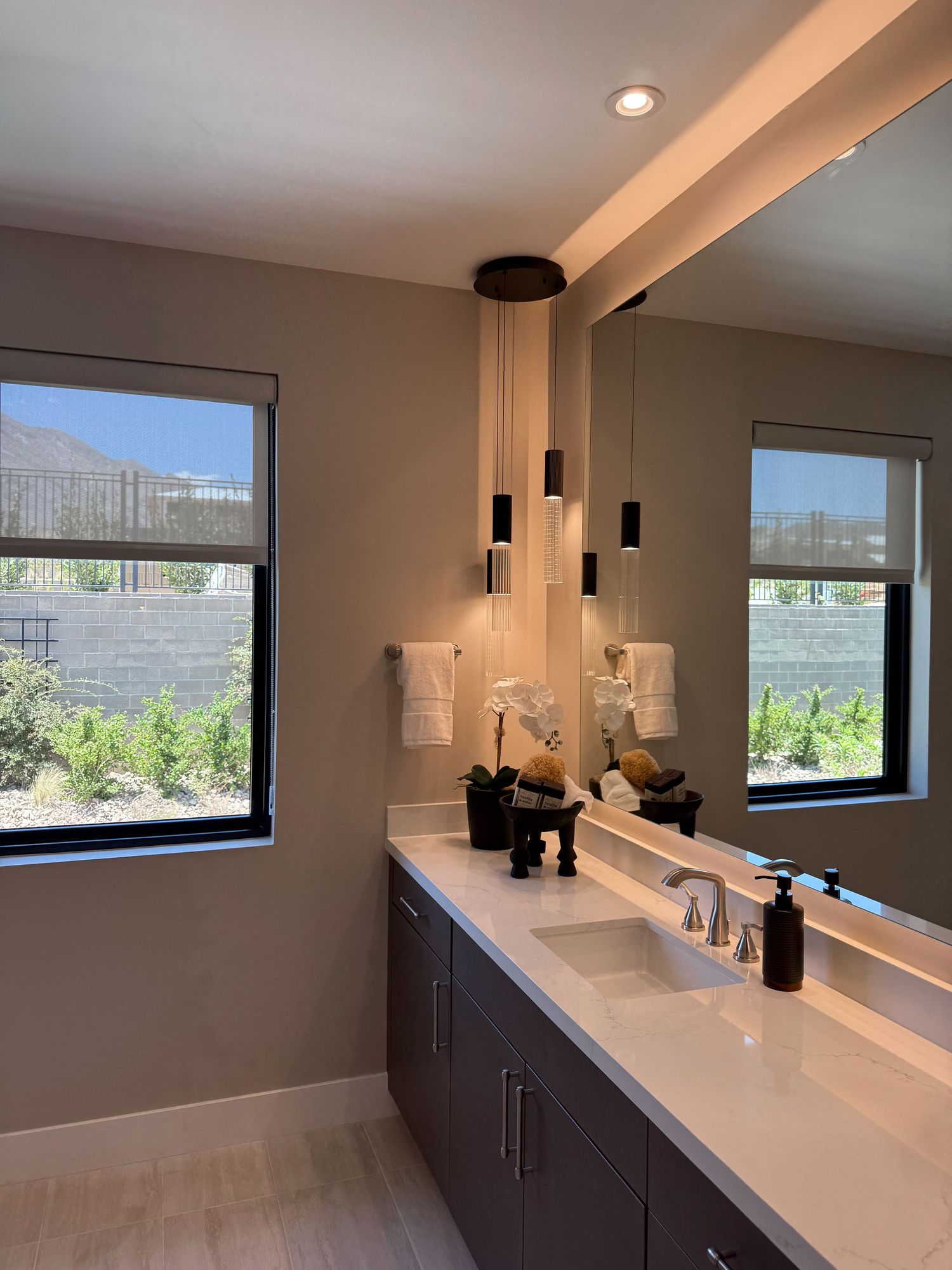Modern bathroom vanity with double sinks, white quartz countertop, dark cabinets, pendant lights, and large mirror with natural light.