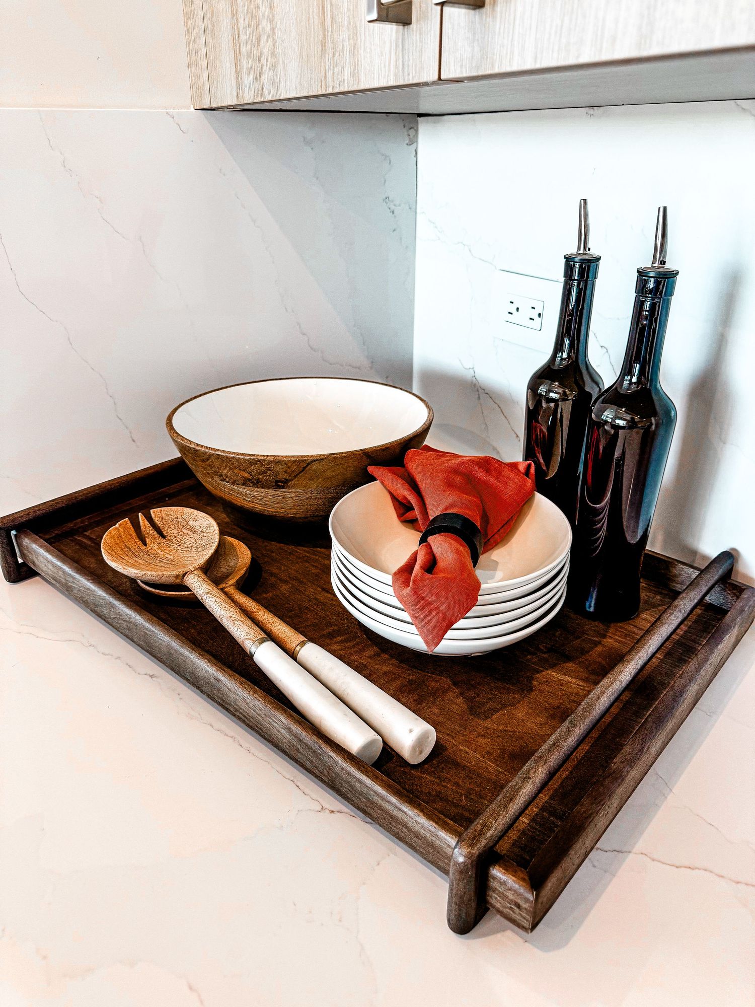 Dark wood serving tray with salad bowl, stacked plates, red napkin, wooden utensils, and dark oil bottles on marble countertop.