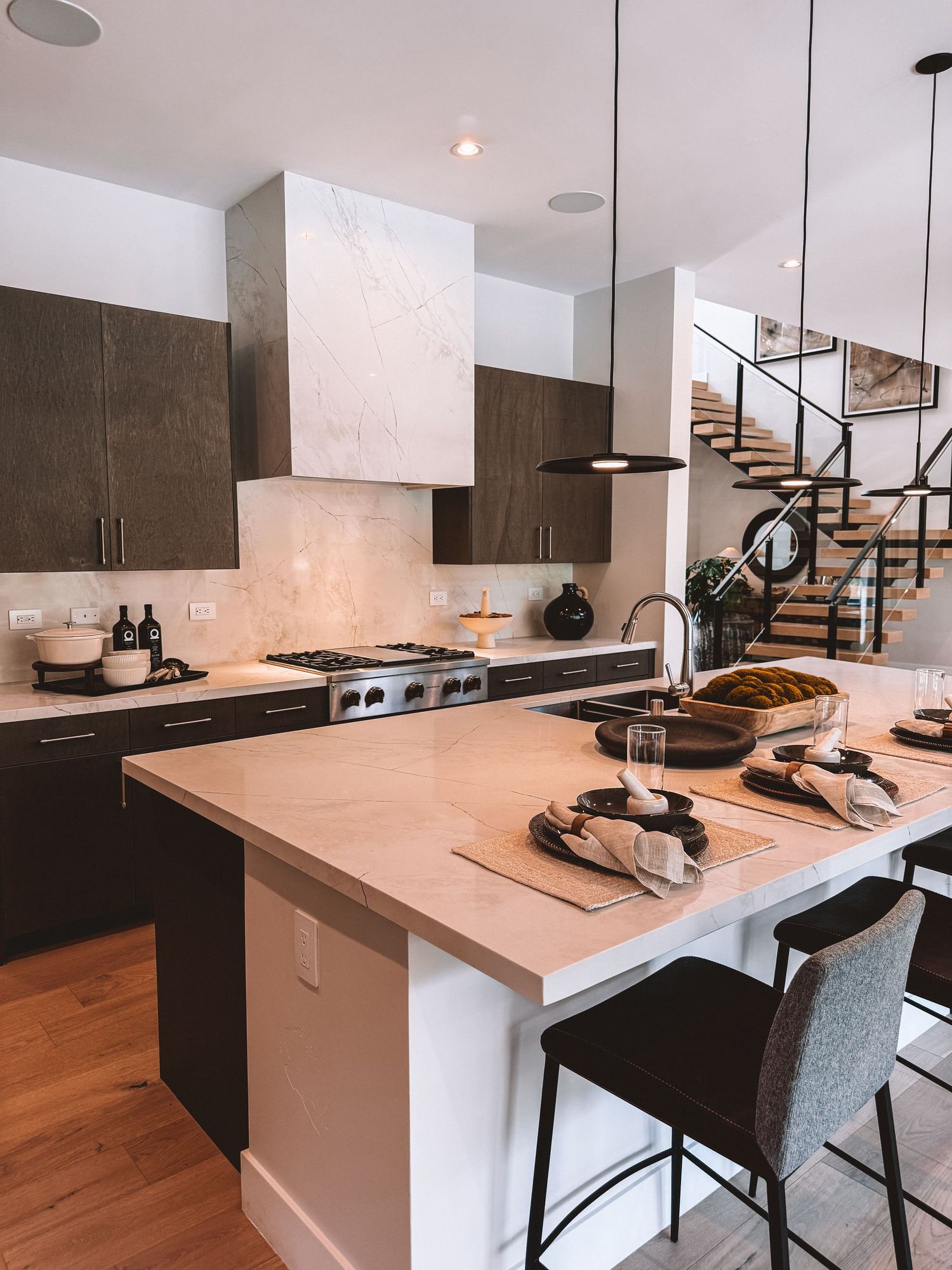 Modern kitchen with dark wood cabinets, marble backsplash, white island, bar stools, pendant lights, and open staircase.