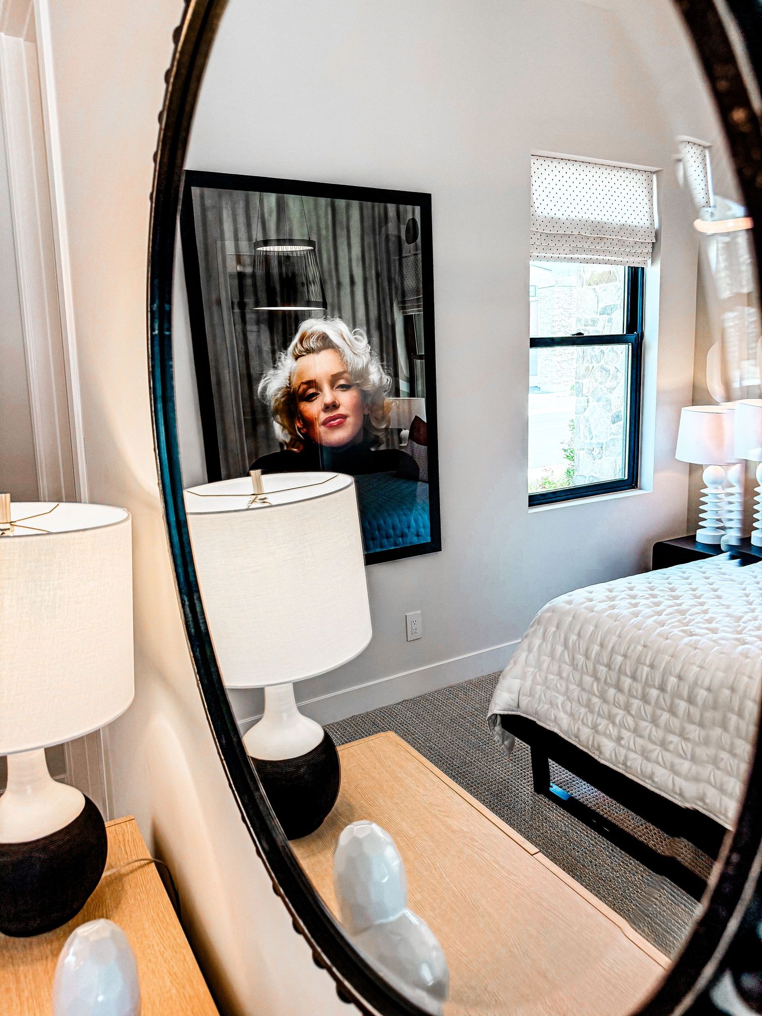 Elegant bedroom reflected in oval black mirror with Marilyn Monroe framed art, white lamps, and neutral bedding.
