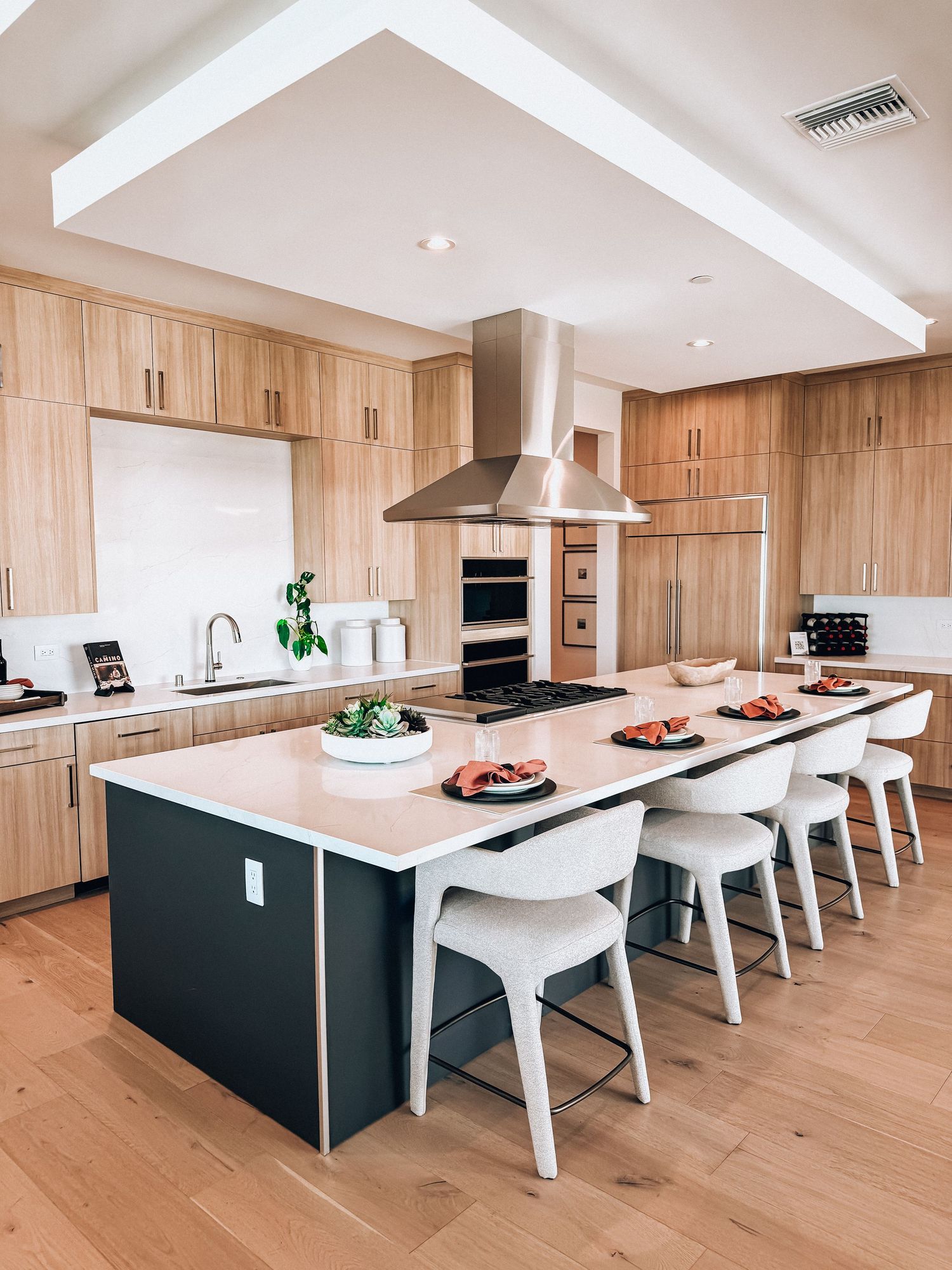 Modern kitchen with light wood cabinets, large dark island, white bar stools, stainless steel range hood, and hardwood floors.