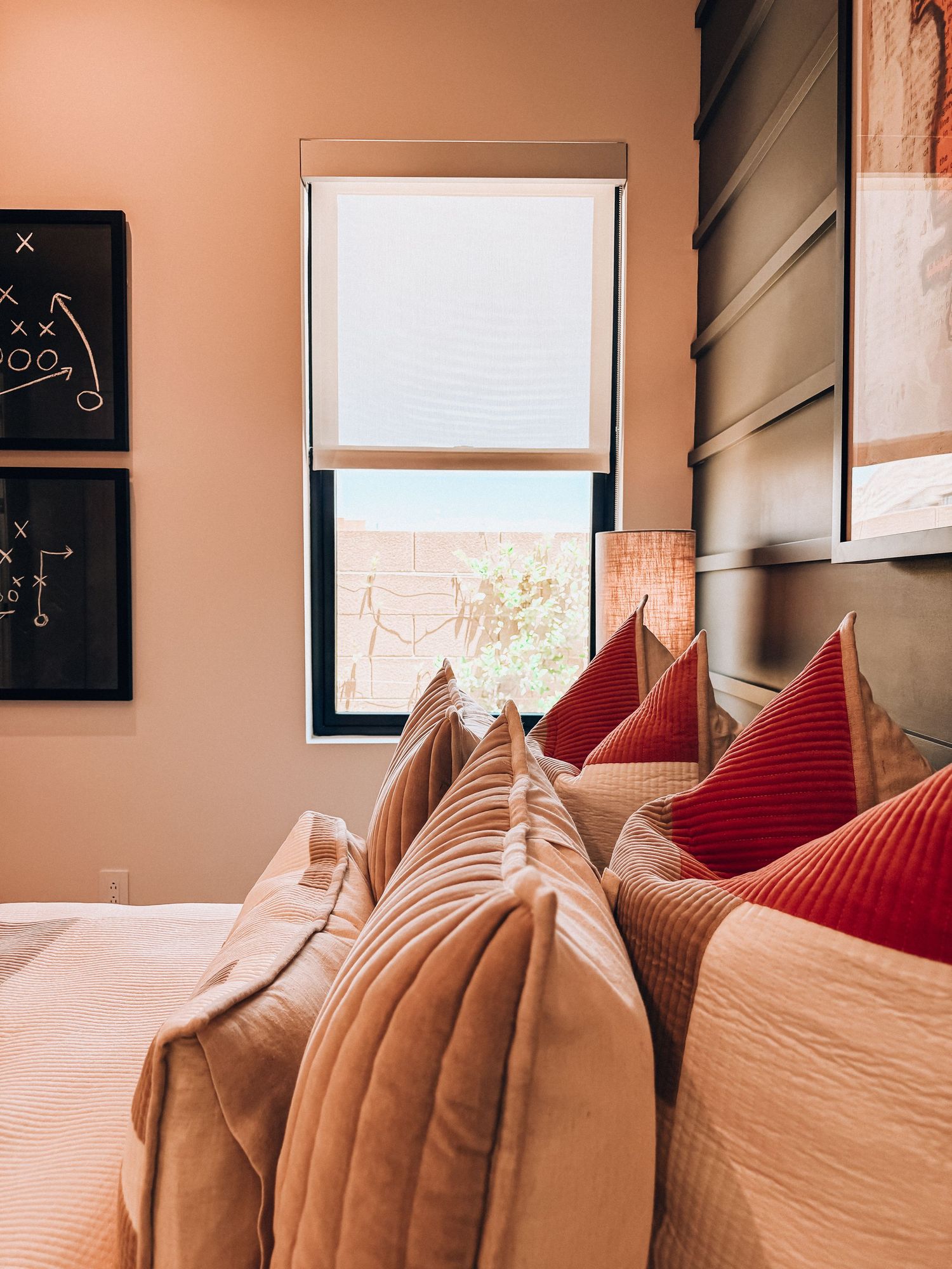 Cozy bedroom with pink and red decorative pillows, dark headboard, roller shade window, and football play art.