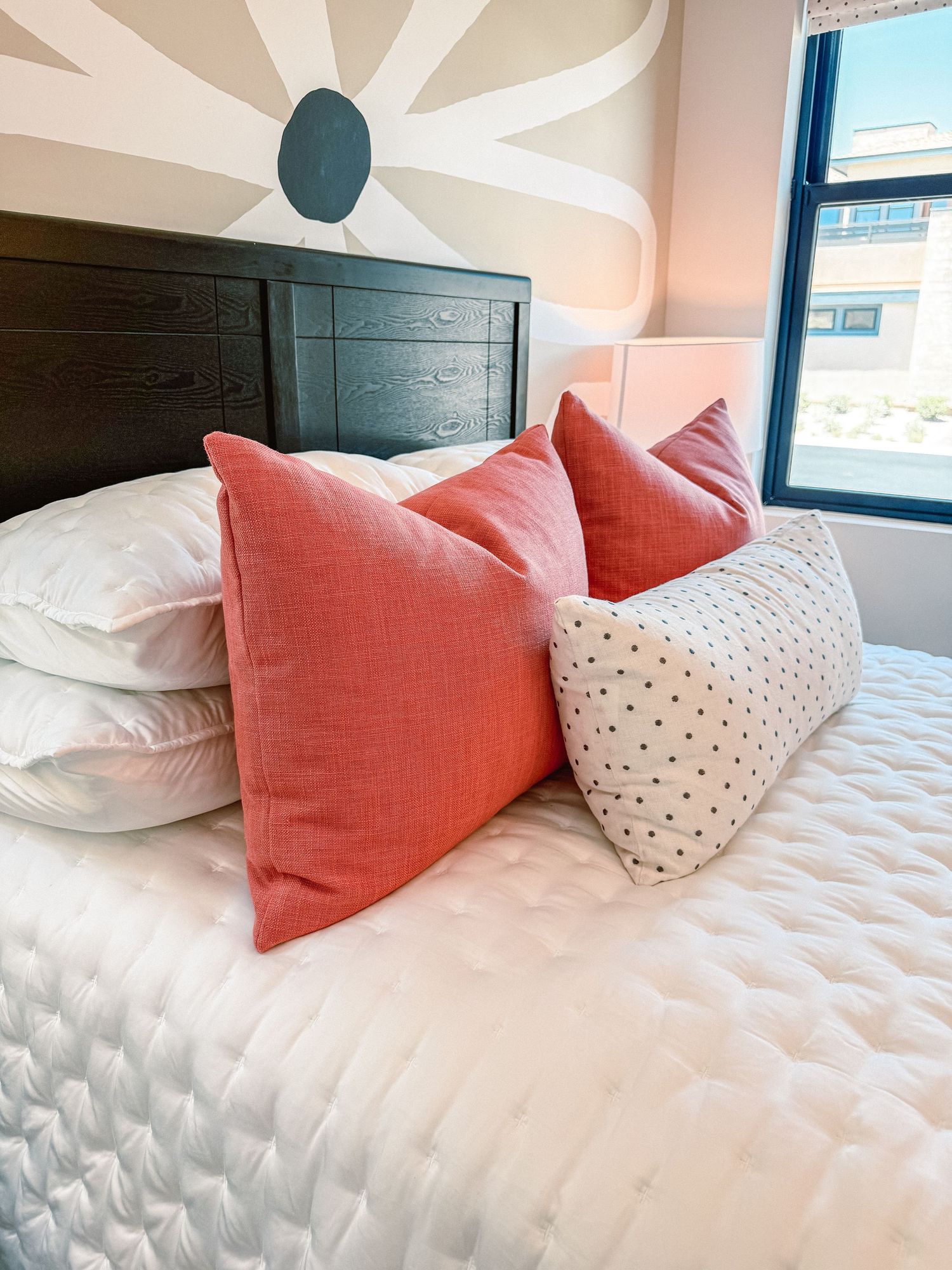 A cozy bedroom bed with coral throw pillows and a white polka dot bolster pillow against a dark wood headboard.