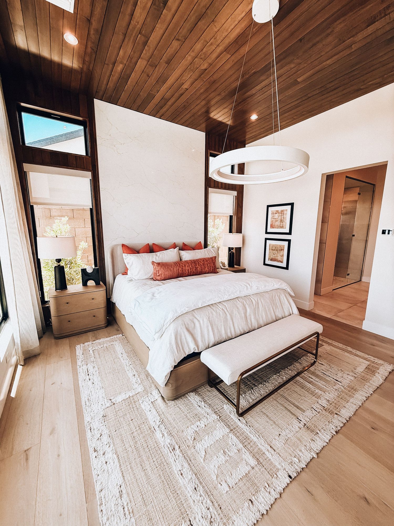 Modern bedroom with wood ceiling, white bedding, terracotta pillows, light hardwood floors, and a circular pendant light.
