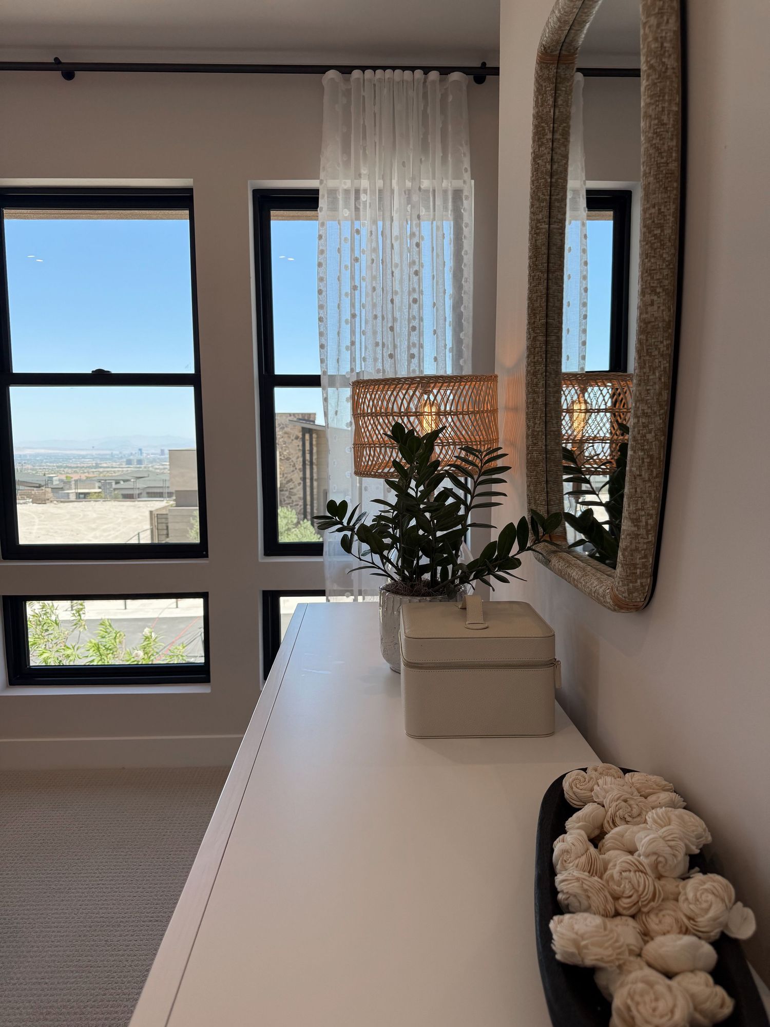 Modern white dresser with plant, mirror, and city view through black-framed windows in a bright bedroom.