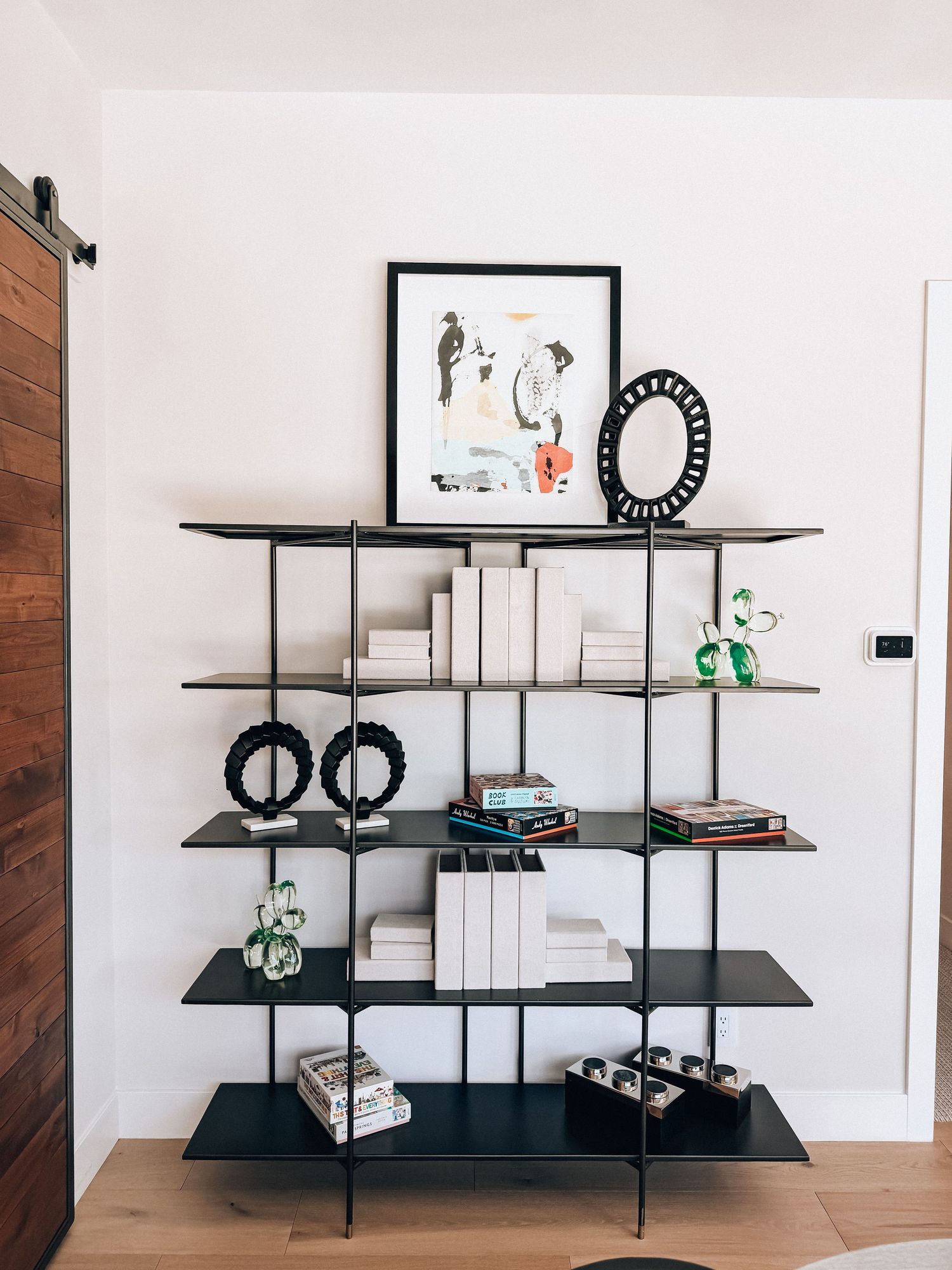 Modern black metal bookshelf with white books, decorative objects, and framed art against a white wall with barn door.