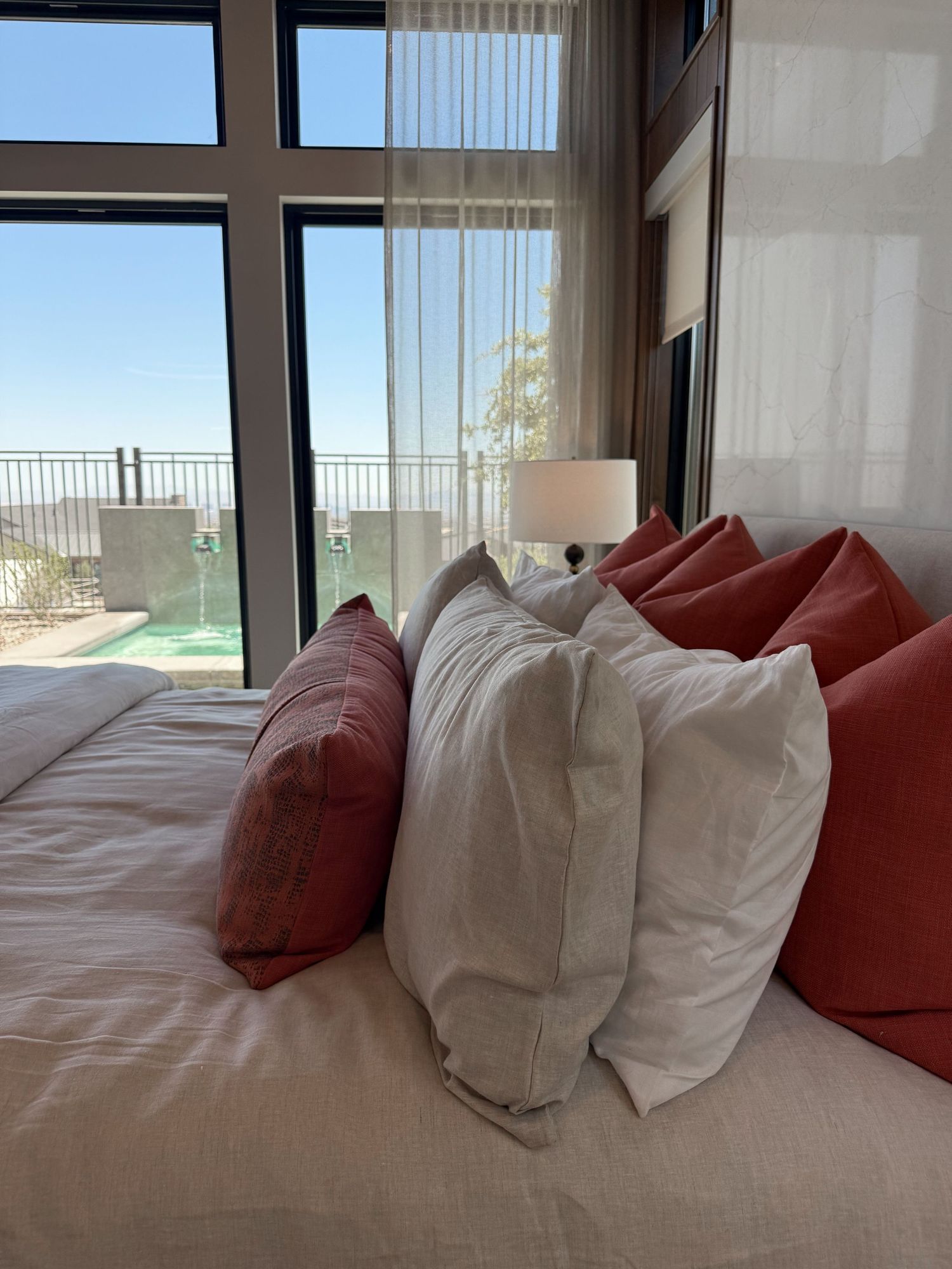 Luxury hotel bedroom with red and white pillows on a bed, featuring floor-to-ceiling windows with ocean view.