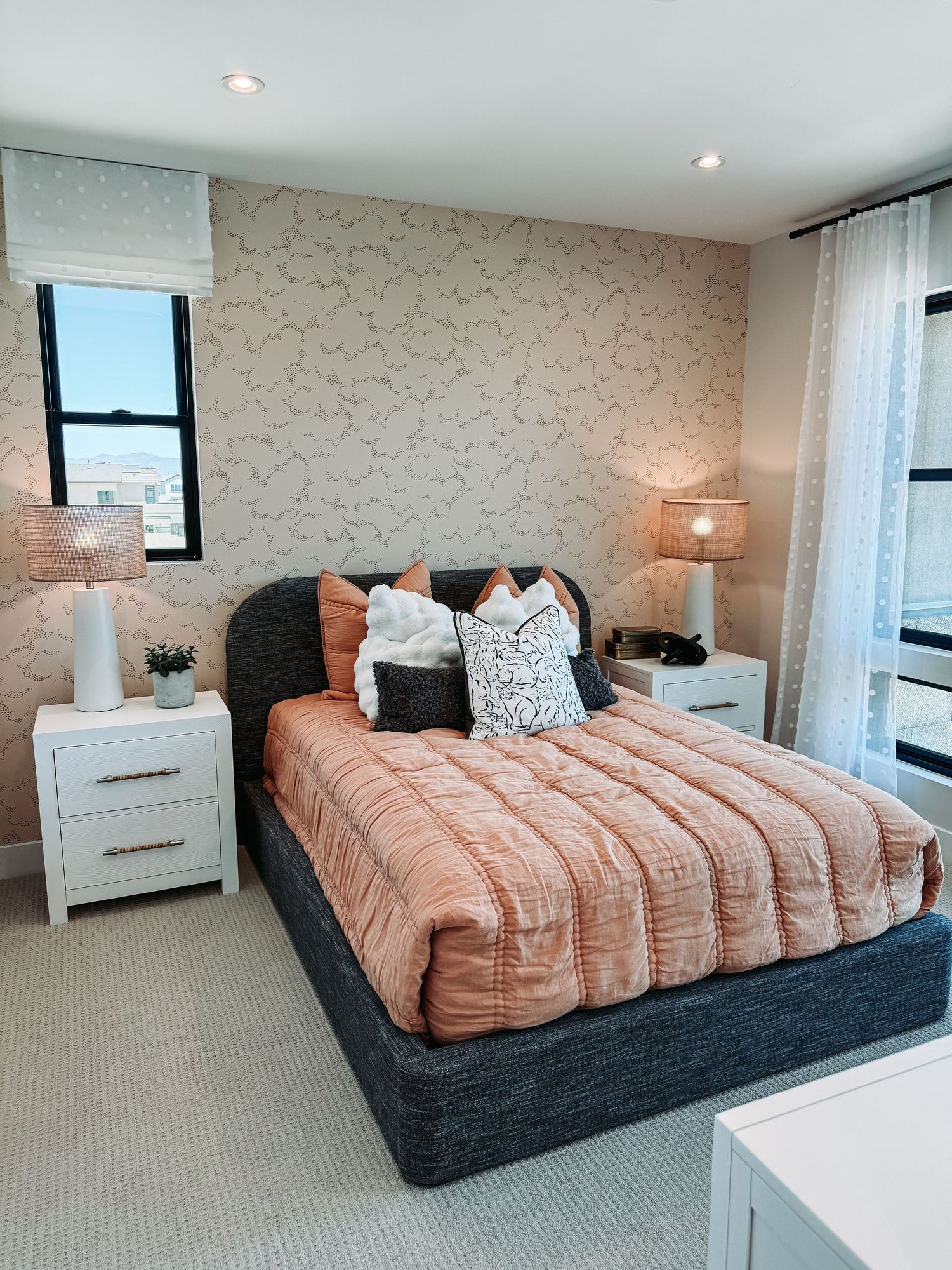 Modern bedroom with peach quilted bedding, dark upholstered bed frame, white nightstands, and floral wallpaper accent wall.