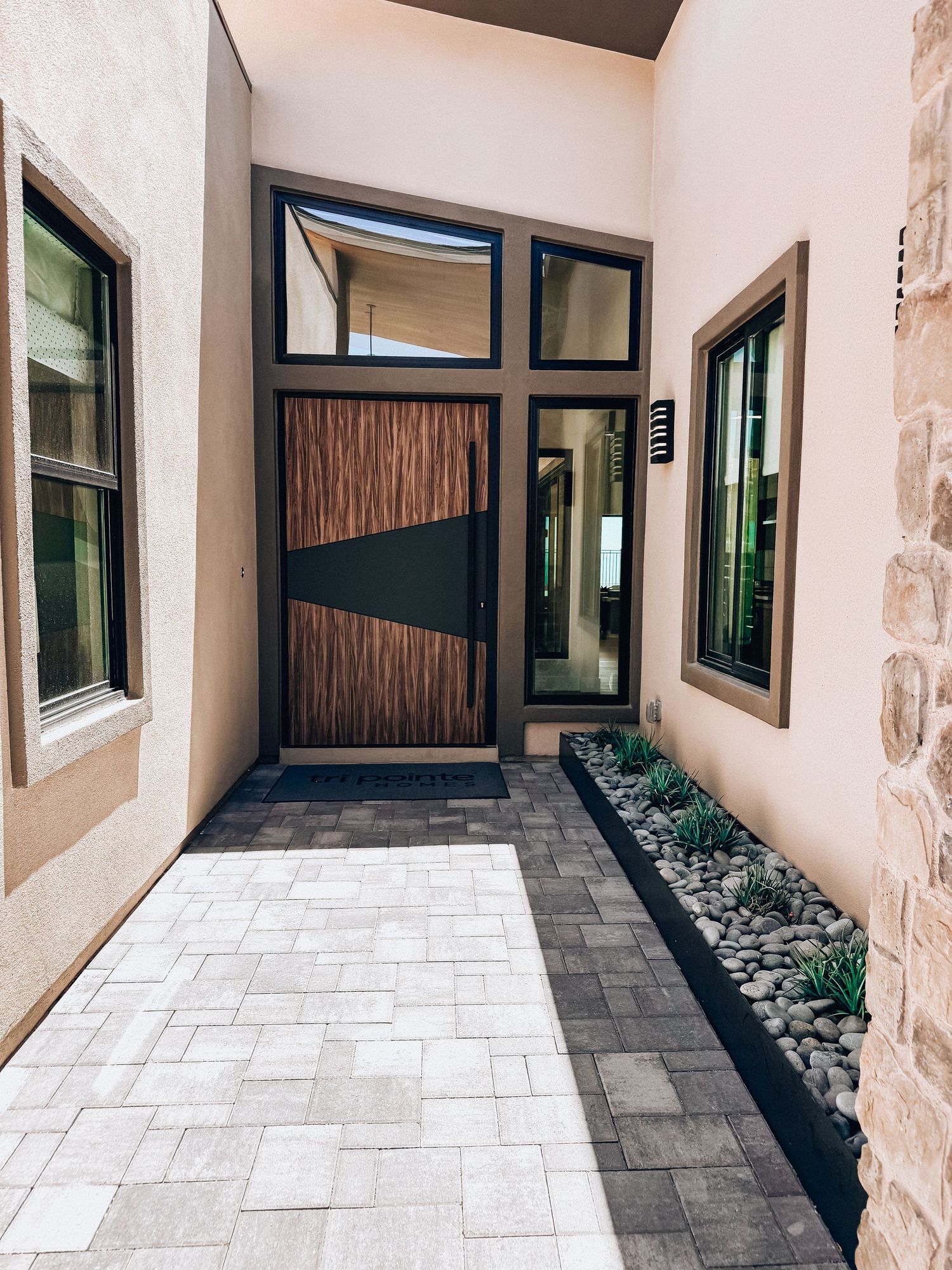 Modern home entrance with wood-textured door, black frame, stone walls, and paver walkway with desert landscaping.
