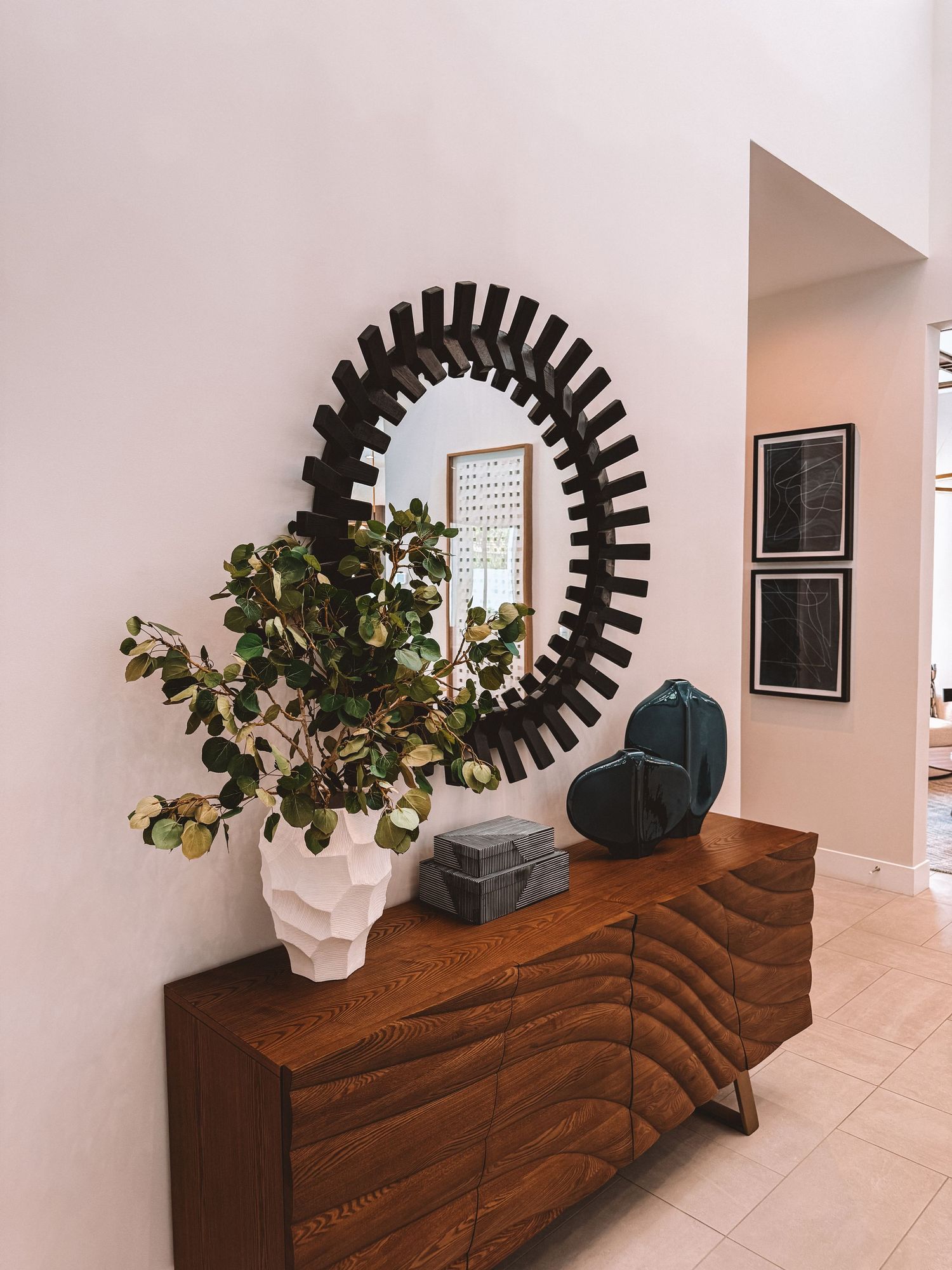Mid-century modern wooden sideboard with decorative mirror, white vase, and teal accents in a bright entryway.