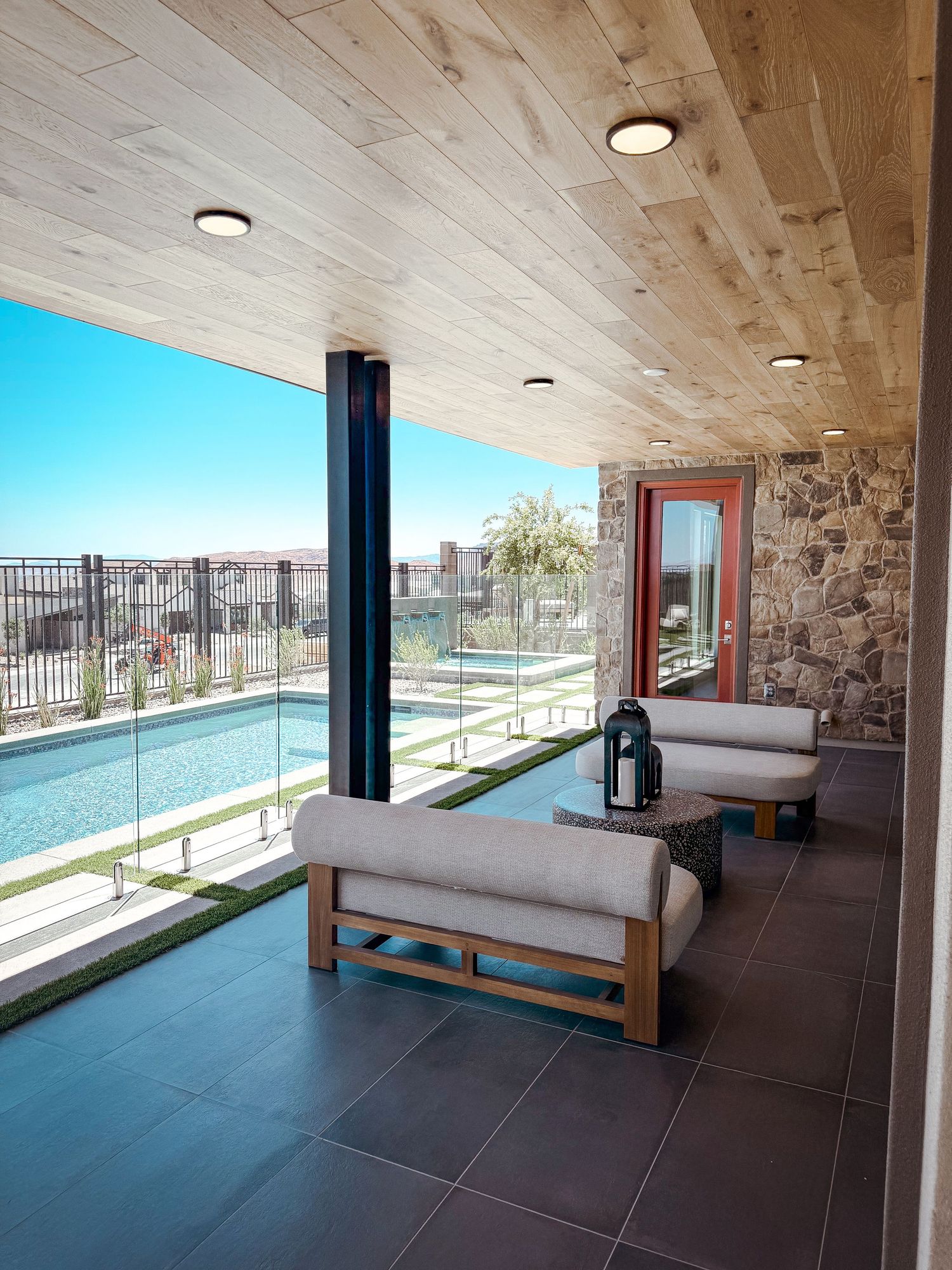 Covered patio with wood ceiling, modern furniture, glass pool view, and stone wall accents in a luxury outdoor living space.