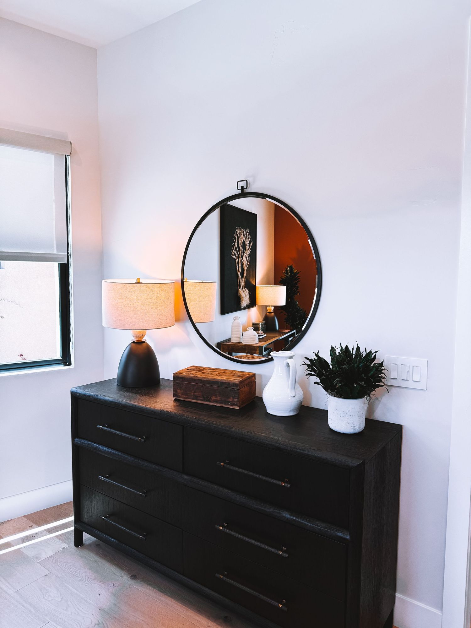 Dark wood dresser with round mirror, lamp, decorative box, and white vase with plant in a modern bedroom.