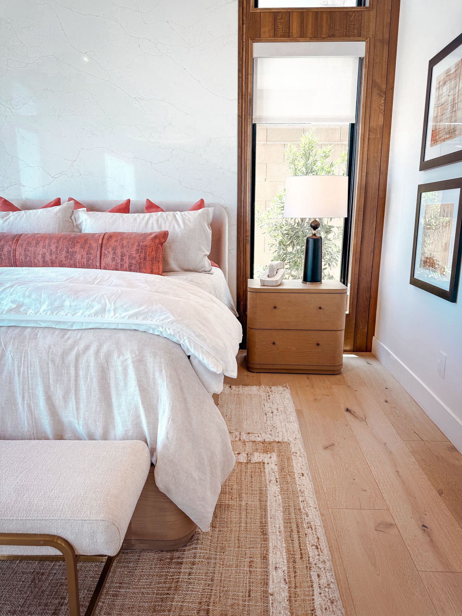 Cozy bedroom with white bedding, coral pillows, light wood floors, a nightstand with lamp, and framed art near a window.
