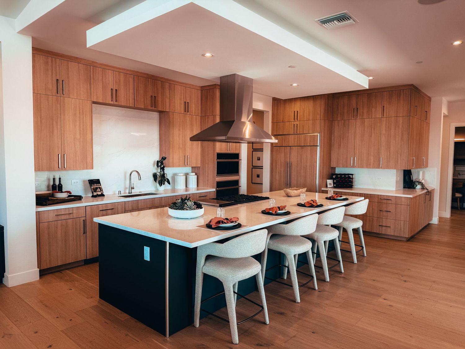 Modern open-concept kitchen with wood cabinets, large island with white chairs, stainless hood vent, and hardwood floors.