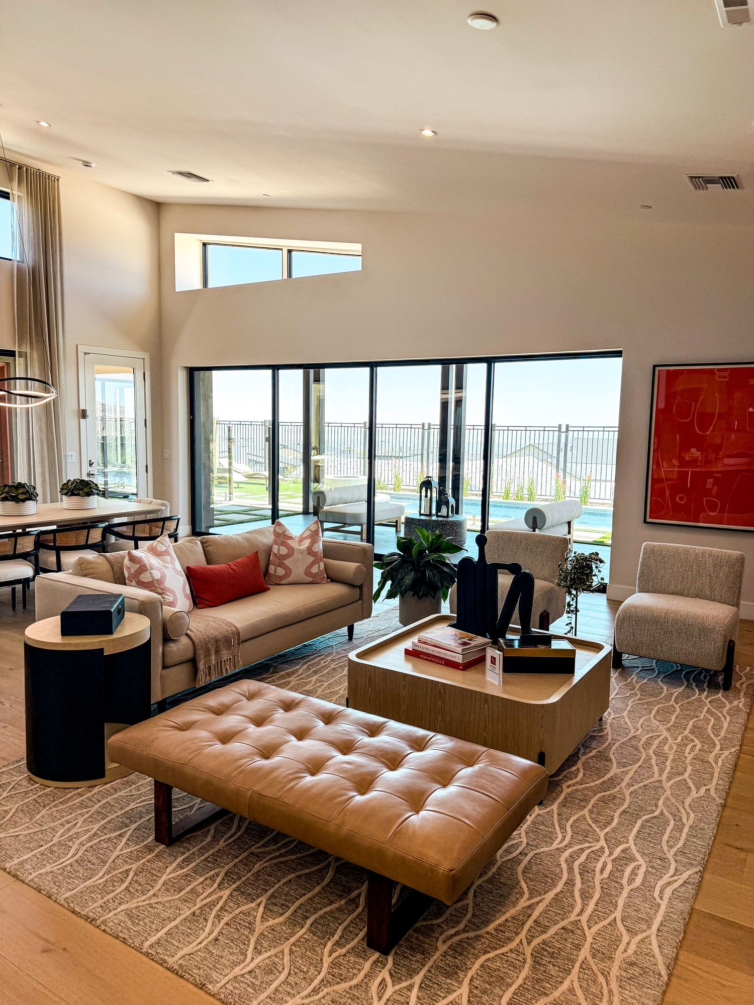 Modern open-concept living room with tufted leather ottoman, neutral sofa, large glass doors, and views of an outdoor pool.