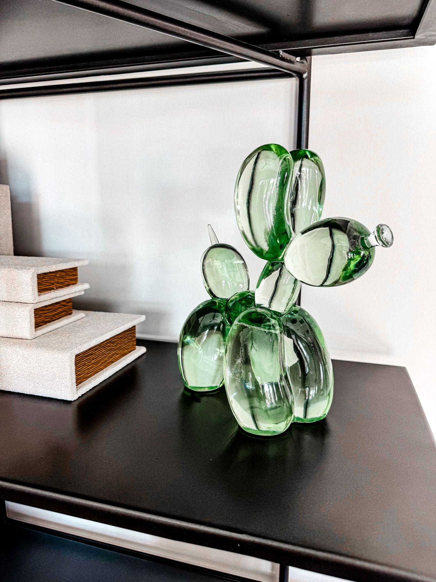 Green glass balloon dog sculpture displayed on a dark shelf beside stacked books in a modern interior.