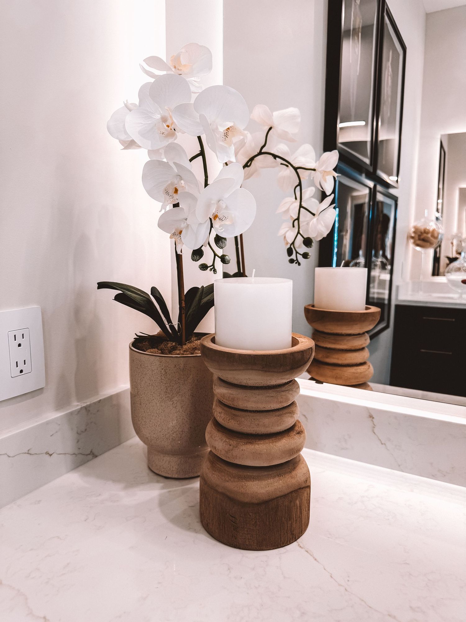 White orchid in brown pot beside wooden candle holders on marble countertop in modern bathroom.