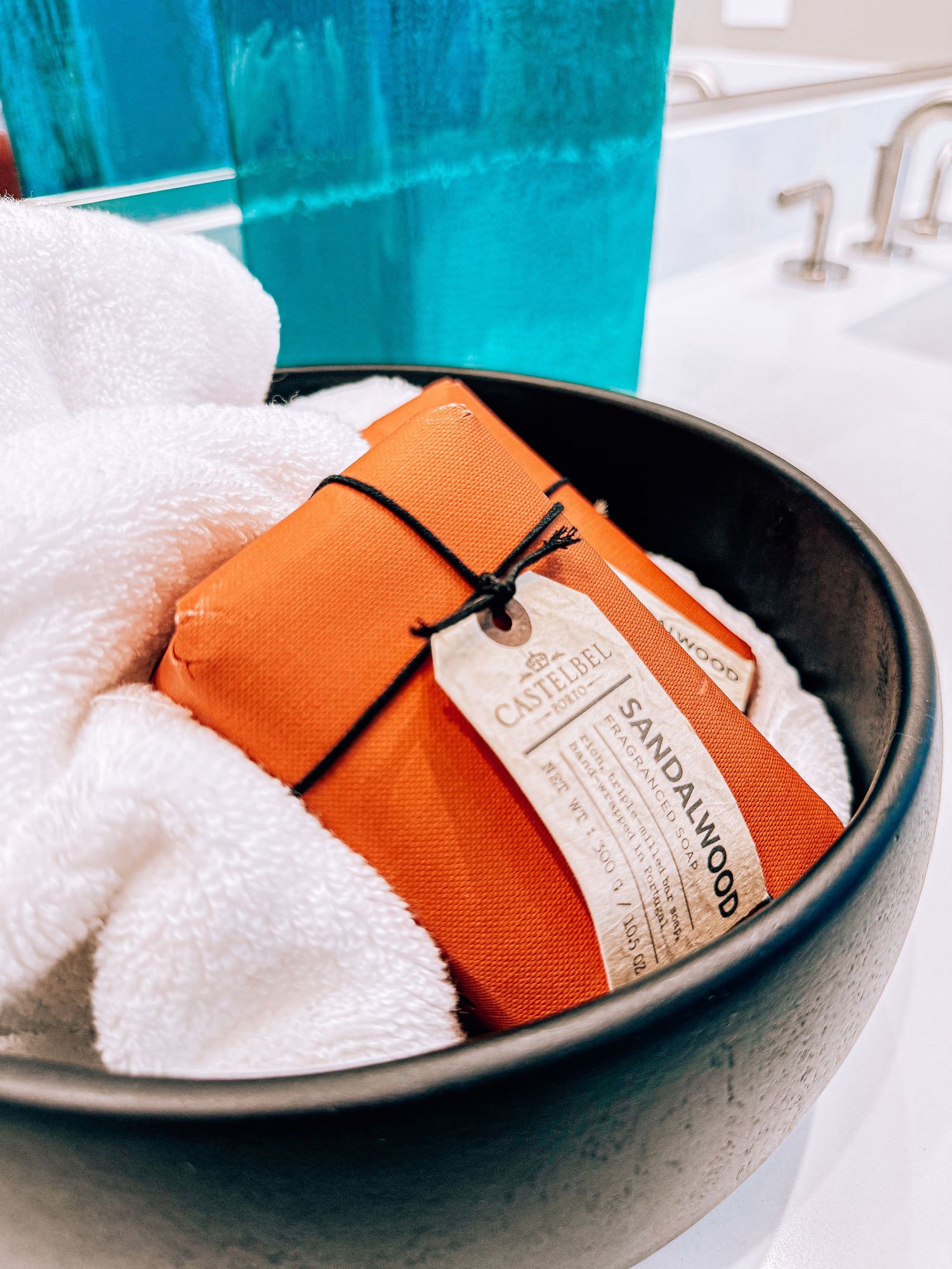 Castelfel sandalwood soap bar in orange packaging nestled in a dark bowl with white towels near a bathroom sink.