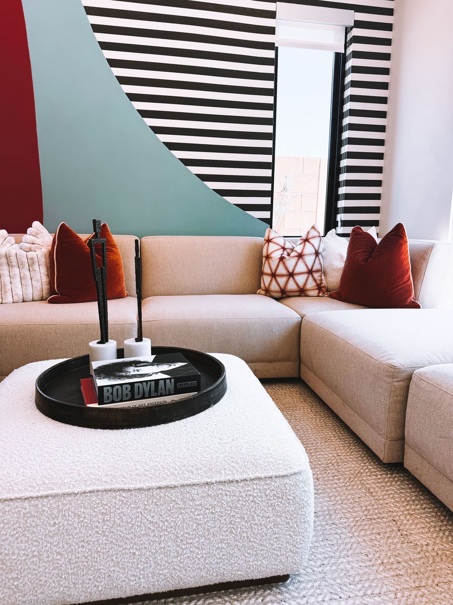 Modern living room with beige sectional sofa, rust throw pillows, white boucle ottoman, and bold black-and-white striped accent wall.