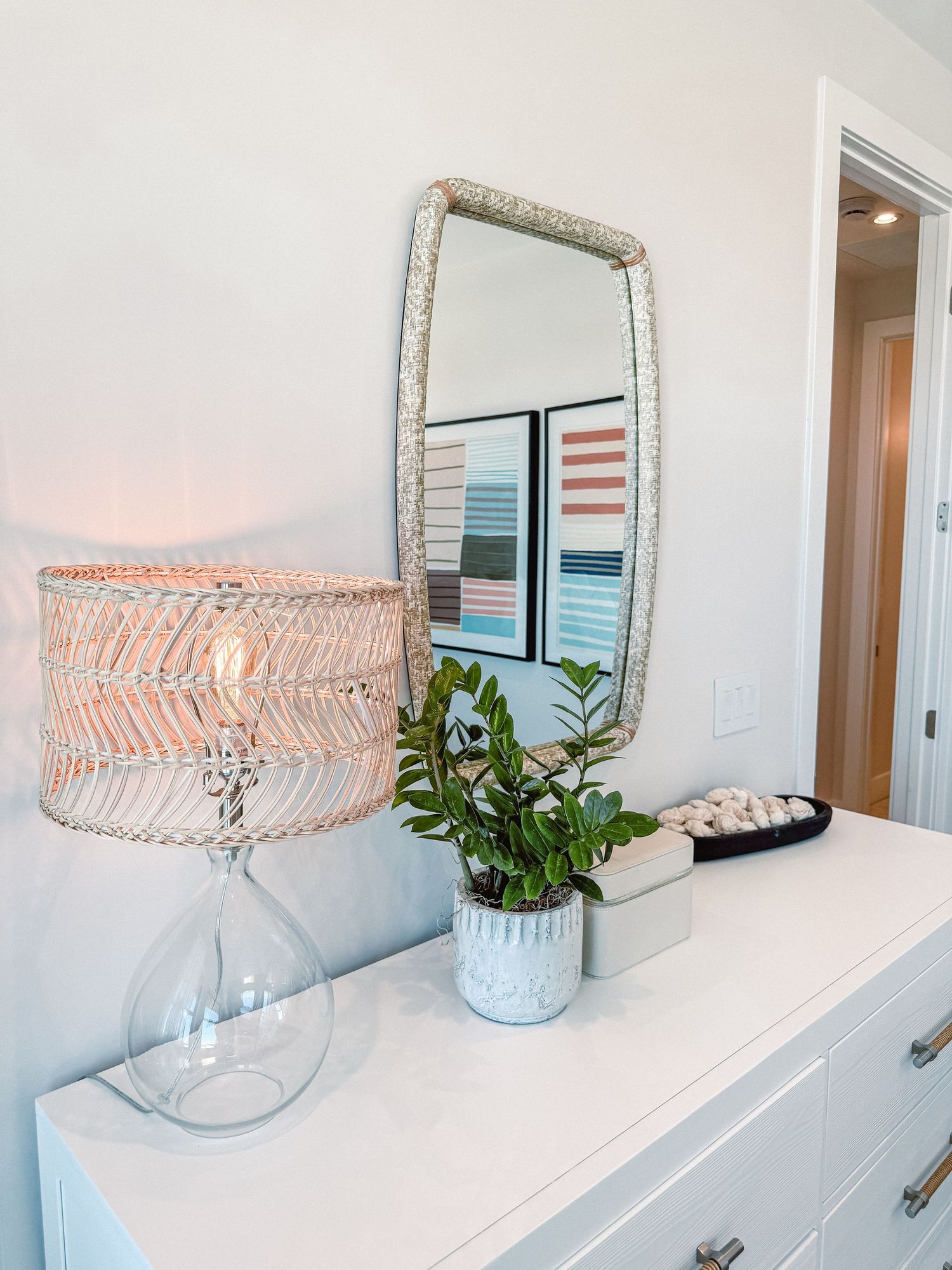 White dresser with woven lamp, green plant, decorative mirror, and abstract art in a bright, modern bedroom.