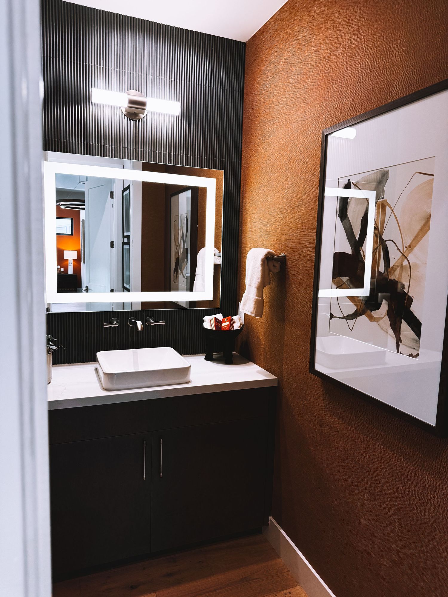 Modern hotel bathroom with dark wood vanity, vessel sink, LED backlit mirror, and abstract wall art on warm brown walls.