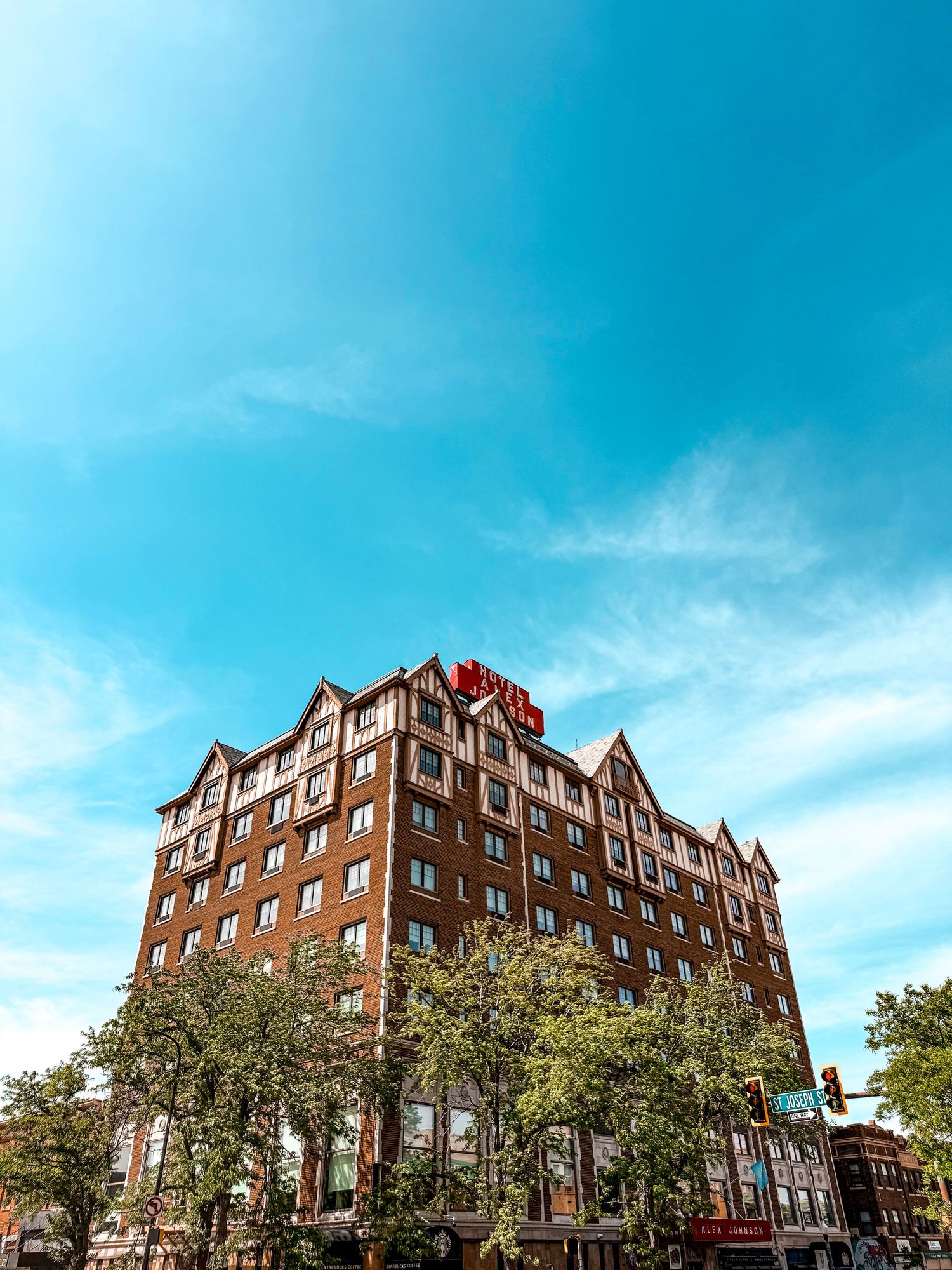 Tall brick hotel building with red rooftop sign surrounded by green trees under a bright blue sky.