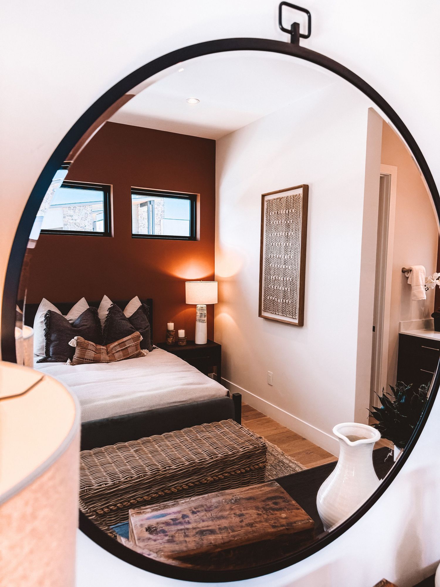 Cozy bedroom reflected in round black-framed mirror with warm brown accent wall, wooden bed frame, and layered textiles.