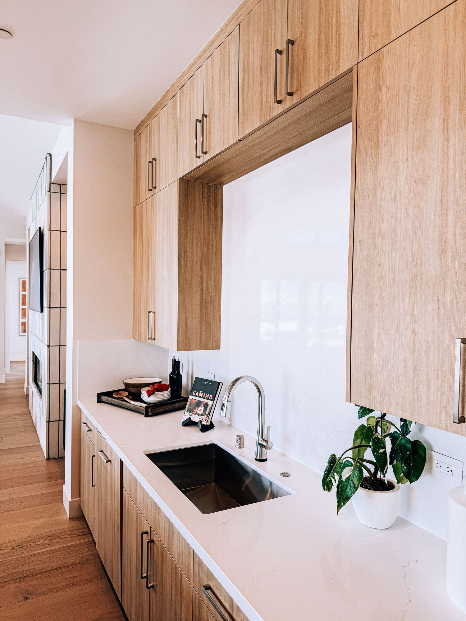 Modern kitchen with light wood cabinets, white quartz countertop, undermount sink, chrome faucet, and a small green plant.